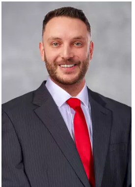 Professional headshot of a man in a dark suit, white shirt, and red tie, smiling against a gray background.
