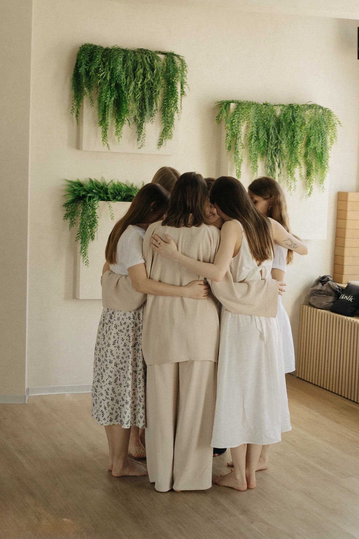Grupo de seis mujeres abrazándose durante un taller de constelaciones grupal en una sala con paredes blancas, decorada con plantas verdes en cuadros en la pared.