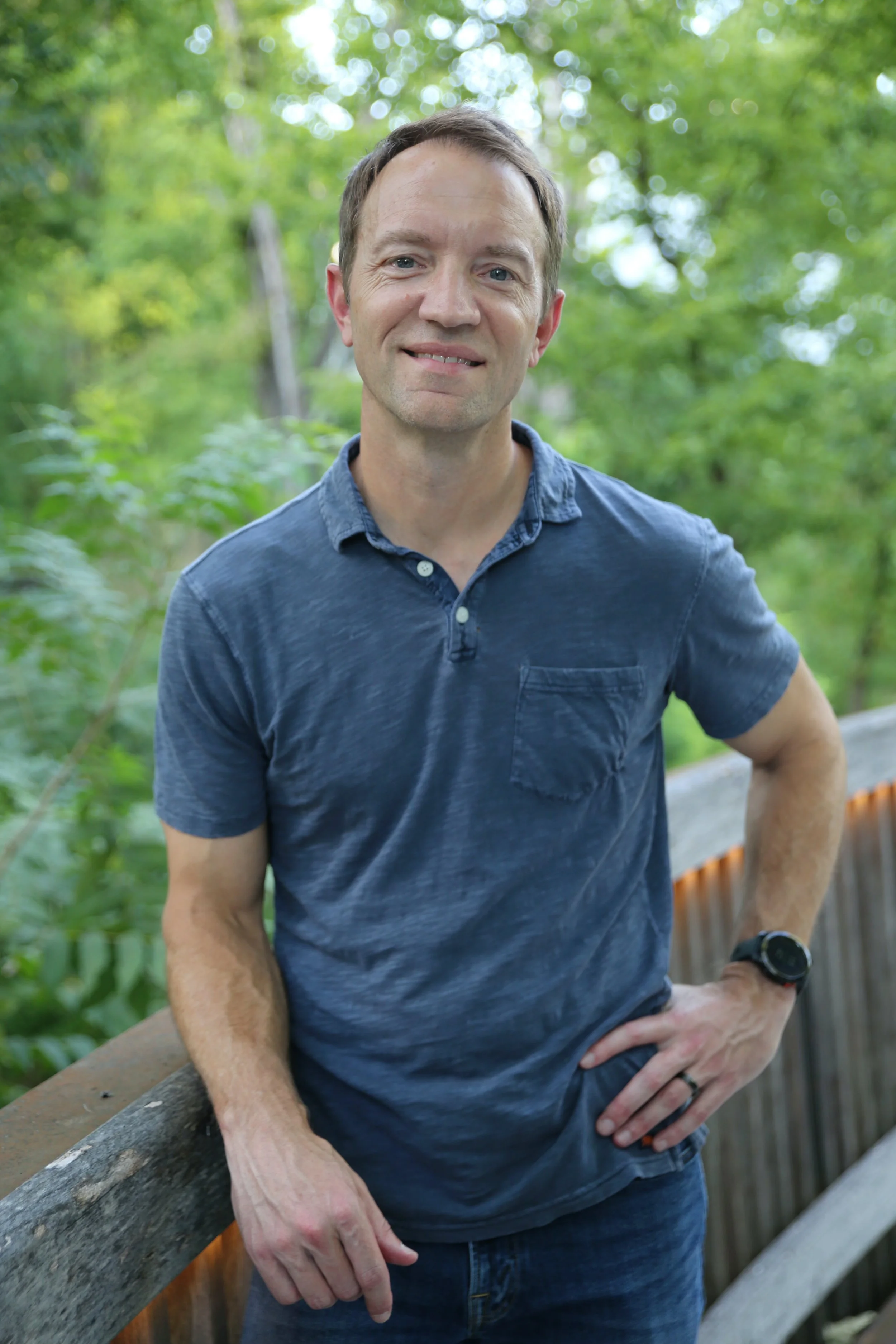 John Coleman, wearing a blue polo shirt and a watch, standing outdoors by a wooden fence with green trees in the background.