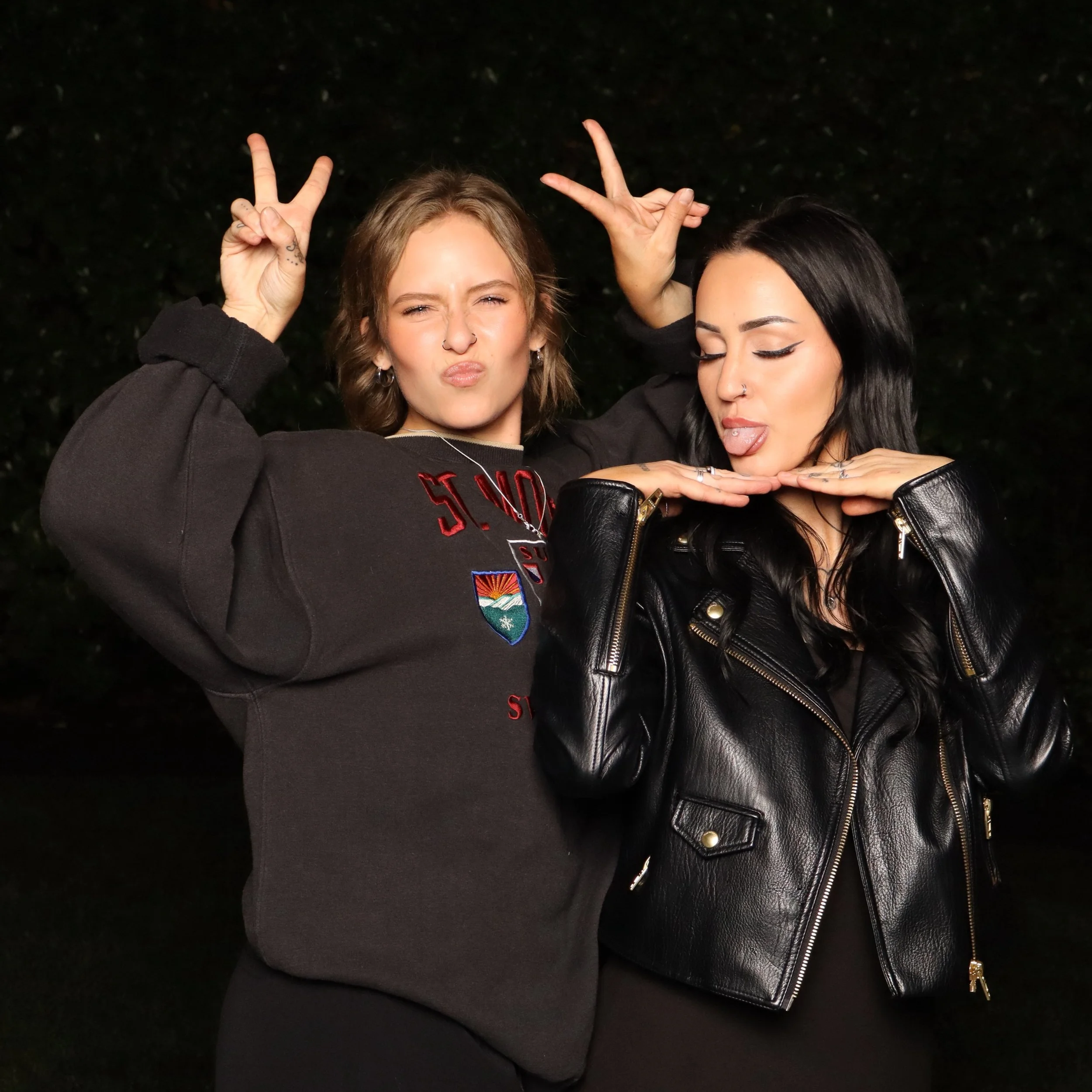 Two women posing outdoors at night, making peace signs with their hands, one with closed eyes and sticking out tongue, wearing black leather jacket and the other with open eyes, puckering lips, wearing earrings and a nose ring.
