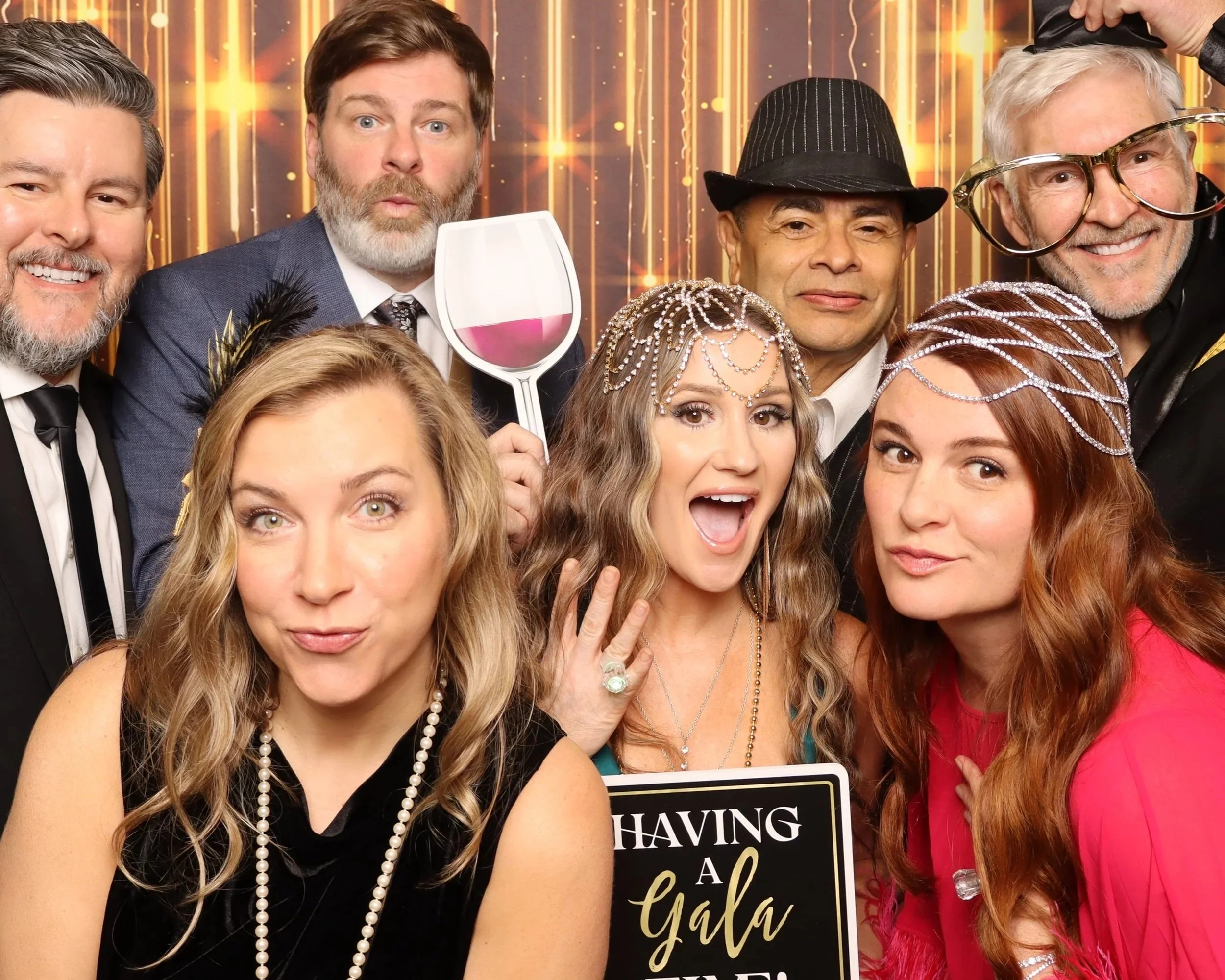 Group of smiling people dressed in fancy attire at a celebration with golden striped backdrop. Some are wearing pearl necklaces, a black hat, and glasses, while one person holds a wine glass and another holds a sign.