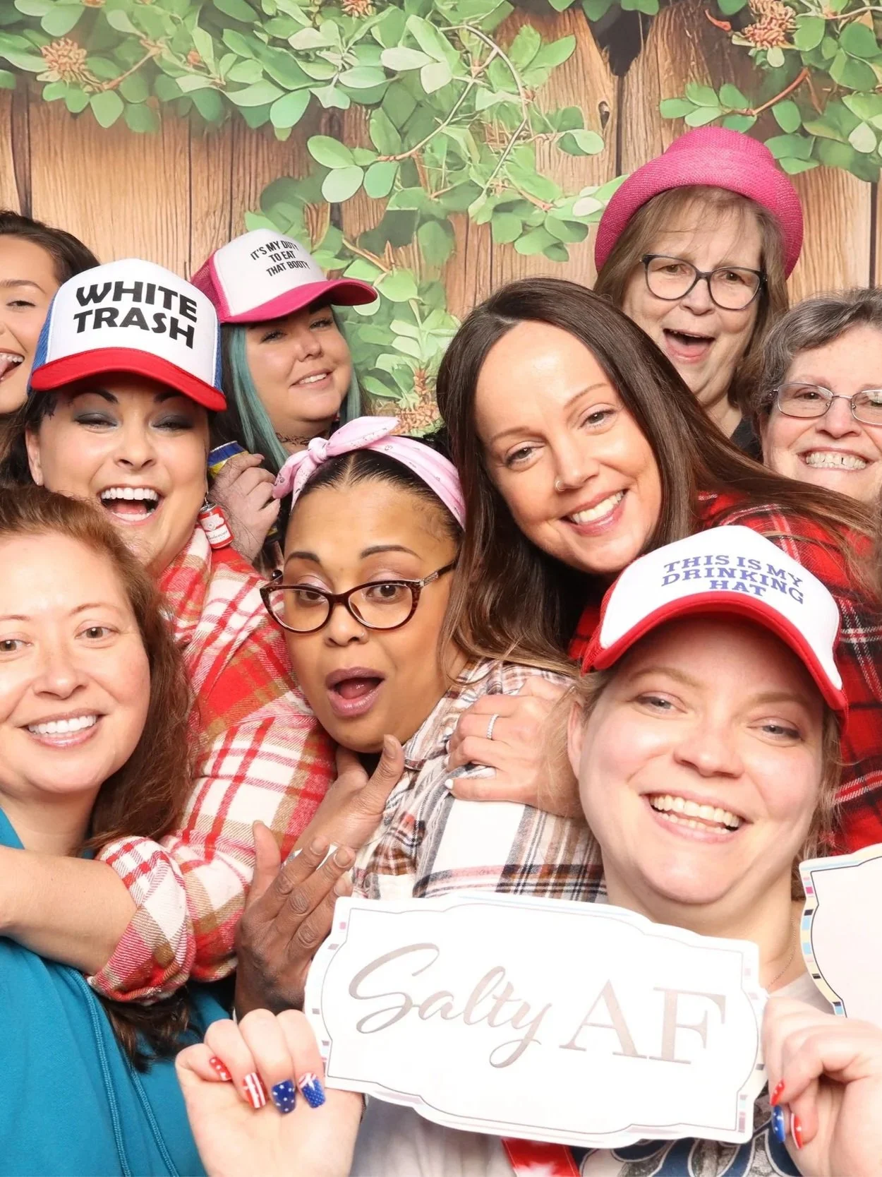 Group of women smiling and posing for a photo, wearing various hats including one with the phrase 'WHITE TRASH' and another with 'THIS IS MY DRINKING HALF'. They are dressed in plaid shirts and are holding signs, with a green leafy background.