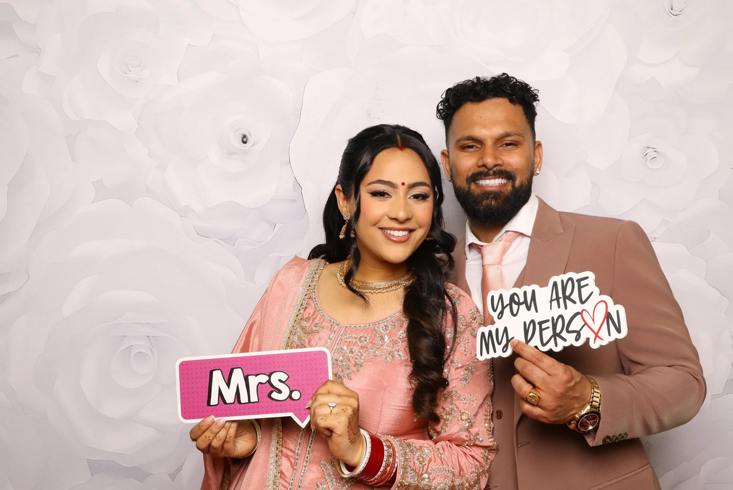 A smiling couple dressed in traditional Indian attire, holding signs that say "Mrs." and "You are my person" with a heart symbol, standing in front of a decorative white floral backdrop.