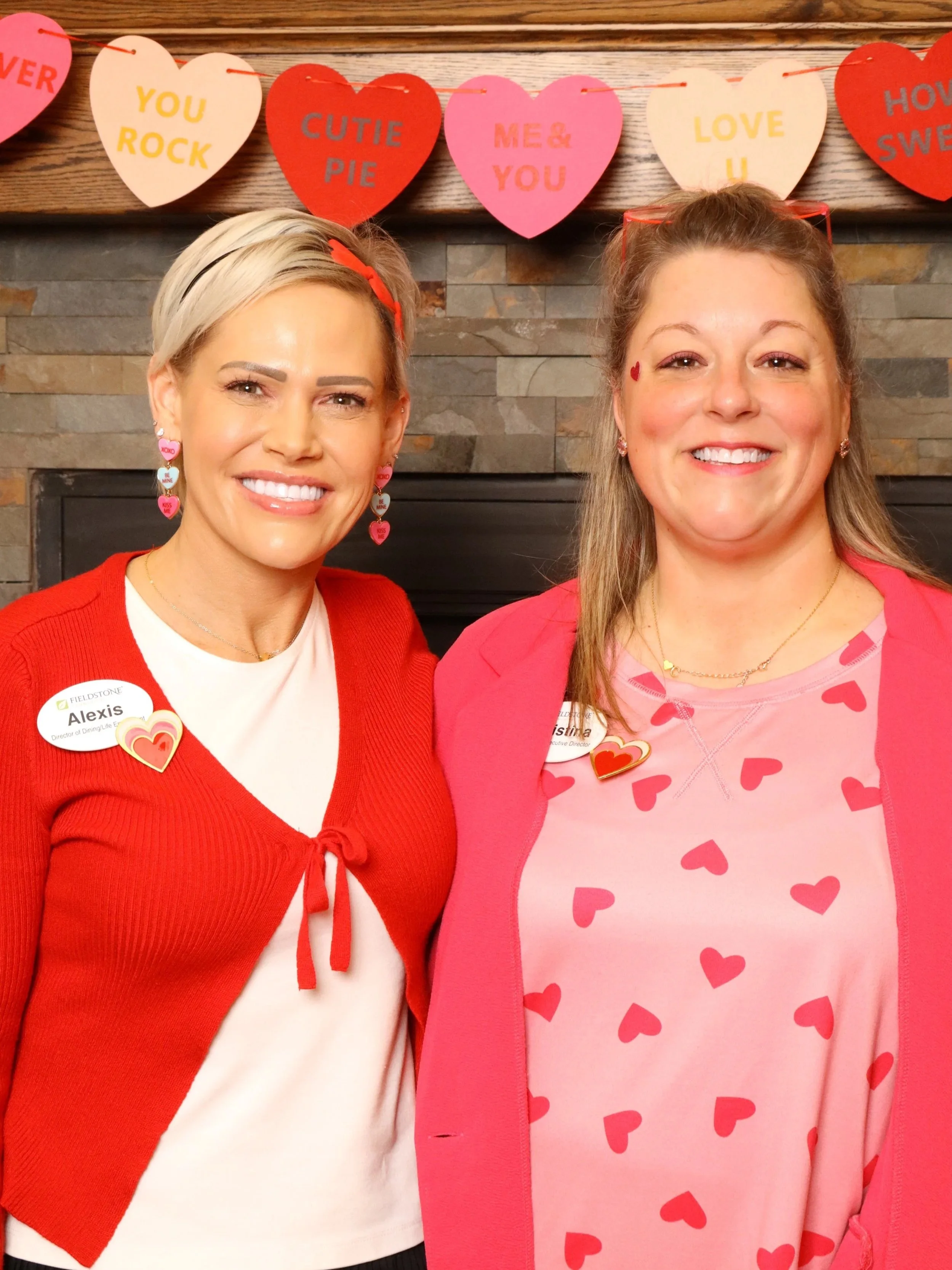 Two women standing in front of a wall decorated with large paper hearts that contain love messages. The women are smiling and wearing red and pink outfits with heart-themed accessories.