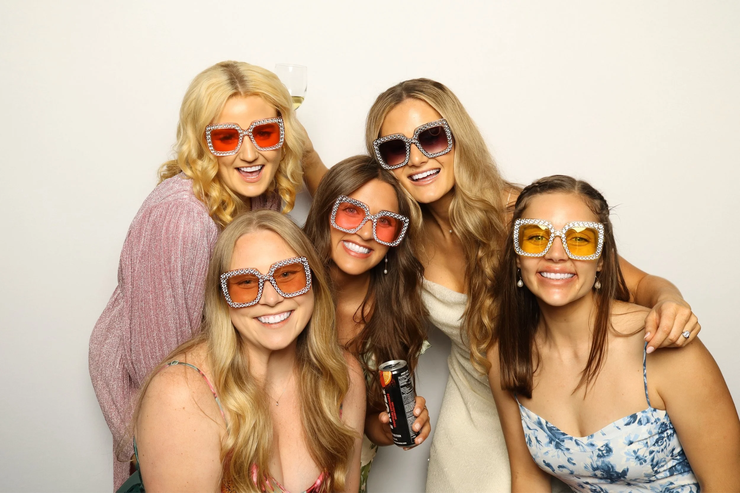 A group of women celebrating together, wearing colorful sunglasses with rhinestones, smiling, and posing for a photo against a plain white wall. One woman is holding a soda can, and one has a glass of white wine balanced on her head.