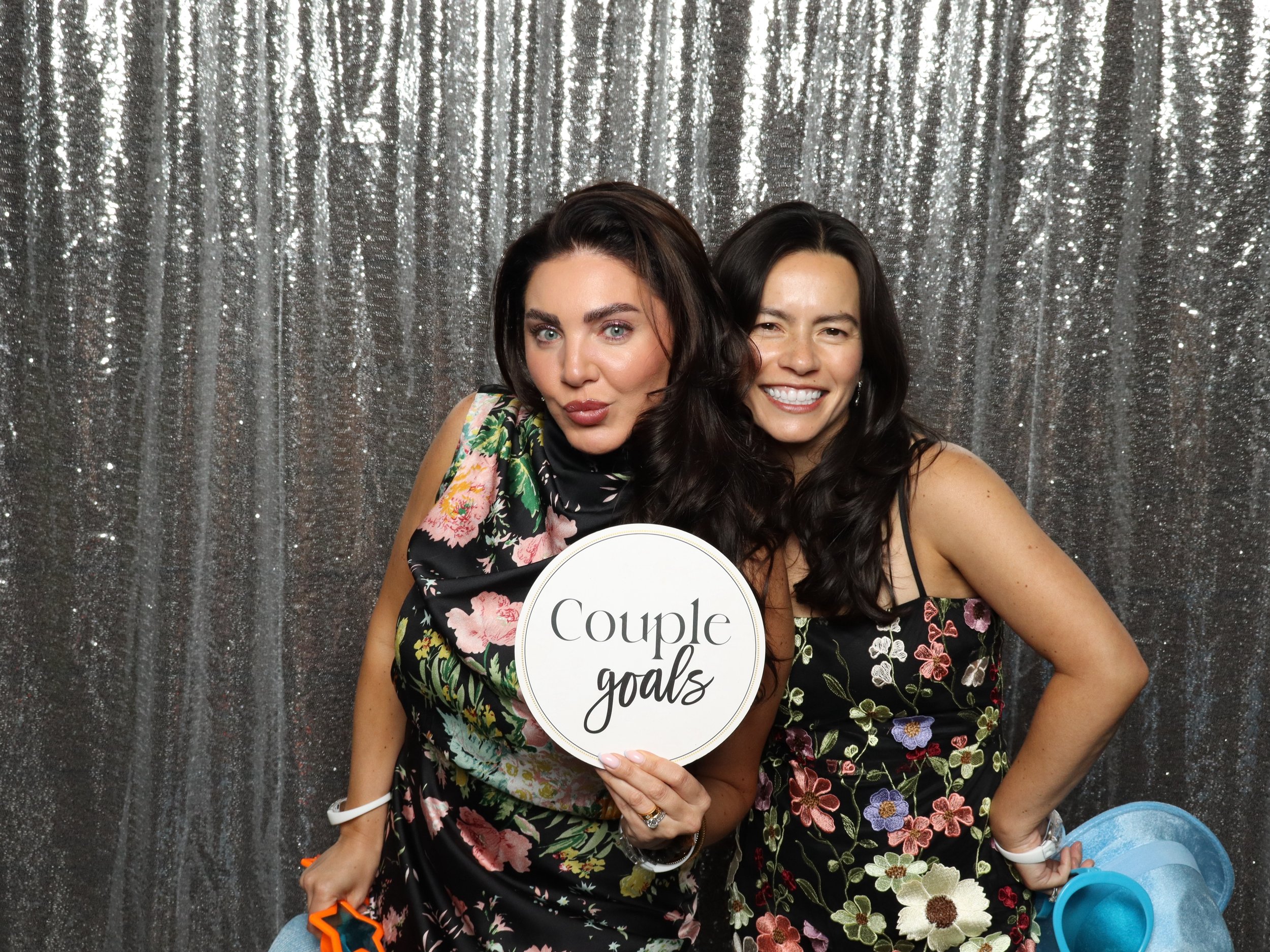 Two women at a photo booth, one holding a sign that reads 'Couple goals,' standing in front of a sparkly silver backdrop.