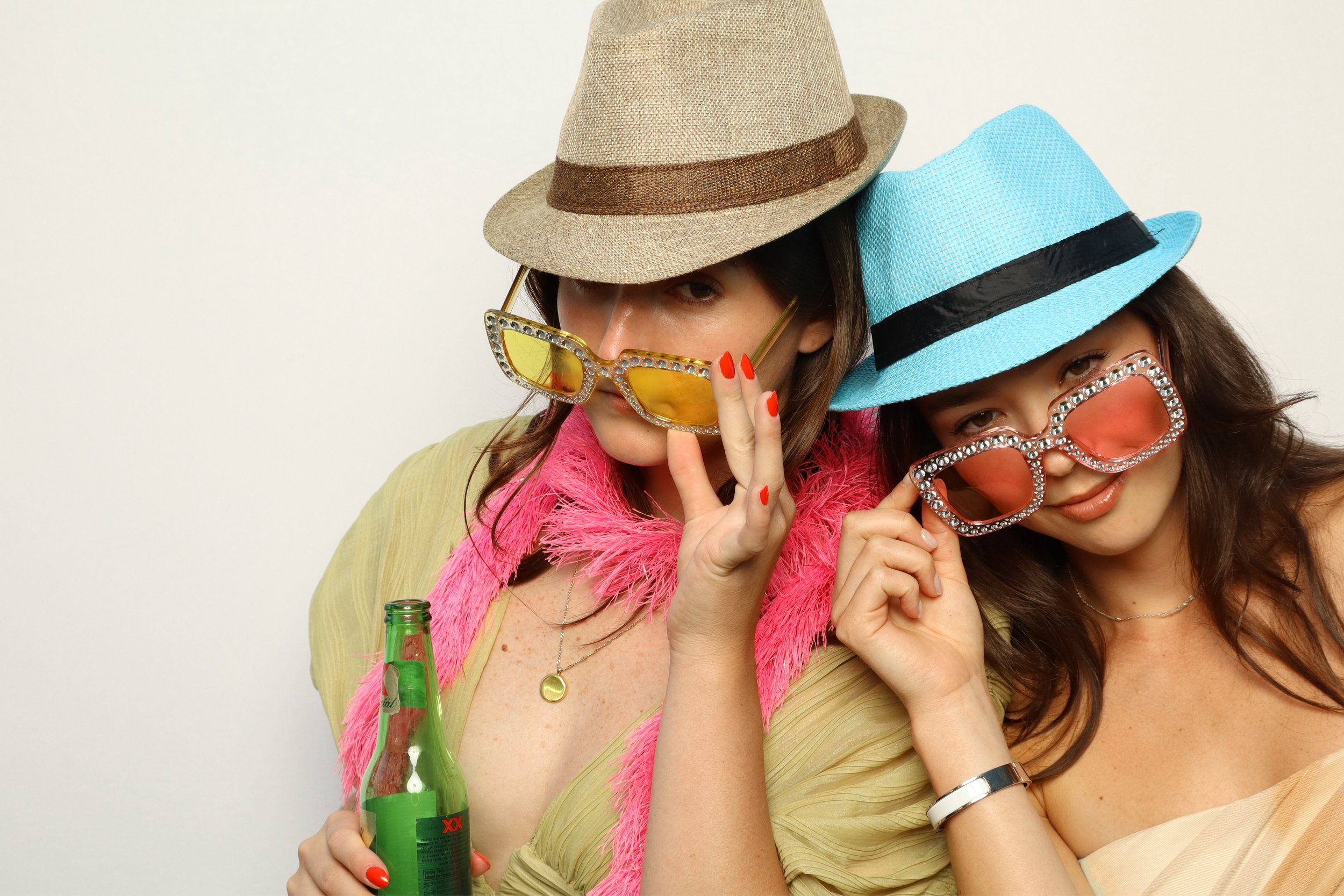 Two women wearing colorful hats and sunglasses, holding a green beer bottle, smiling and posing for the camera in a casual setting.