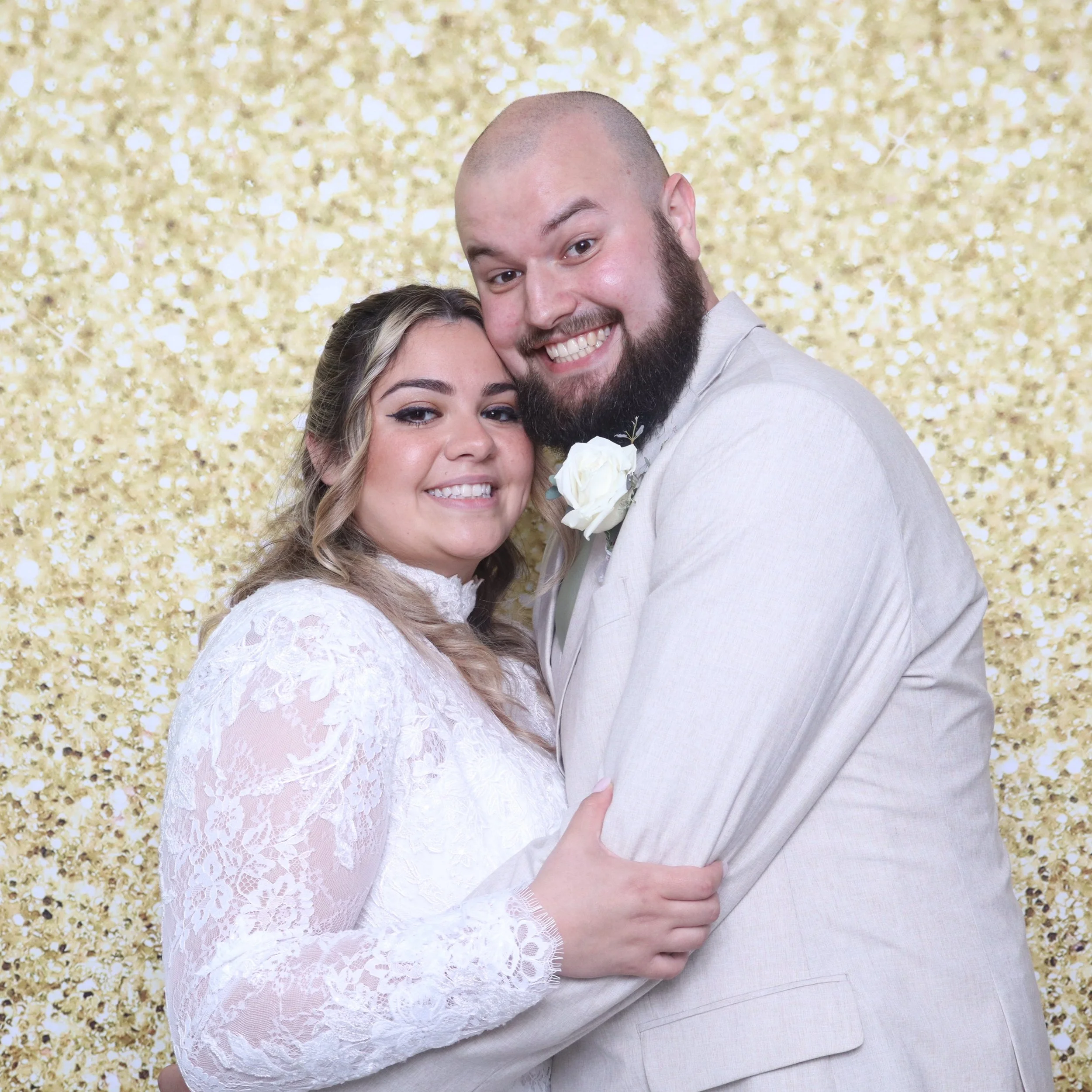 A couple smiling in front of a gold glitter backdrop, with the woman in a white lace dress and the man in a light-colored suit.