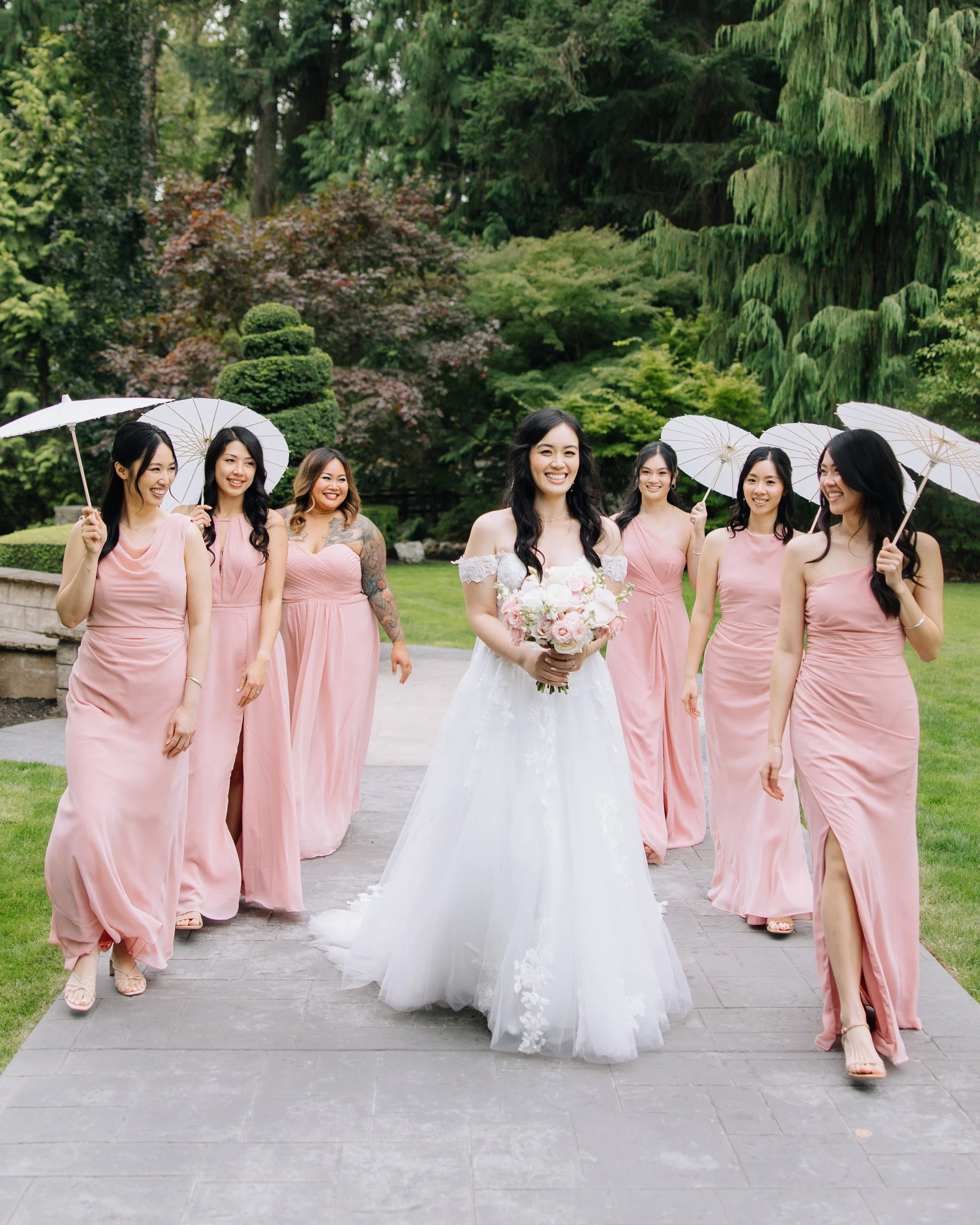 Bride walking with bridesmaids in blush dresses and parasols through a garden setting.