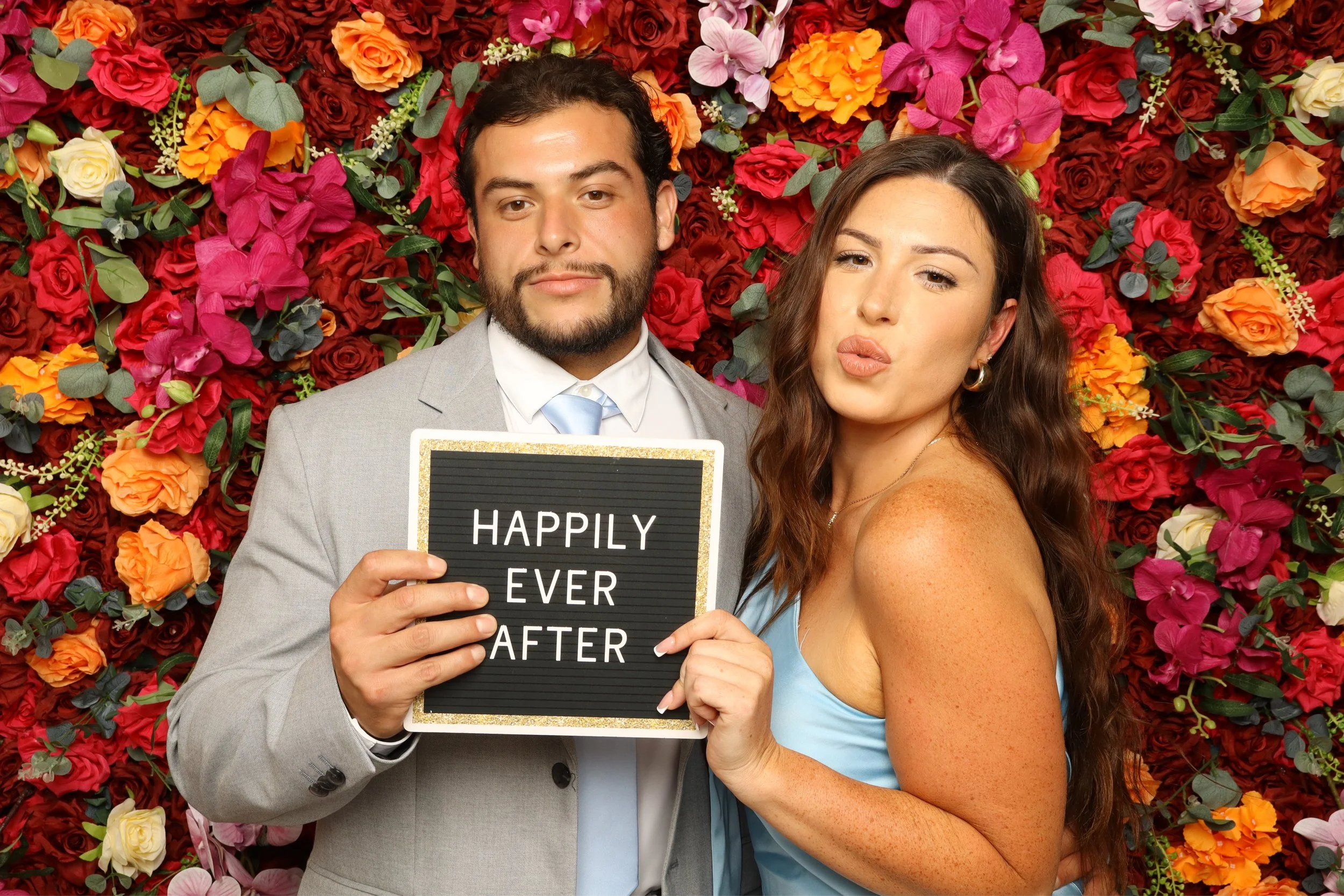 A couple taking a selfie in front of a colorful flower wall, the man in a gray suit holding a sign that reads 'HAPPILY EVER AFTER'. The woman has long brown hair and is wearing a light blue dress, making a kiss face.