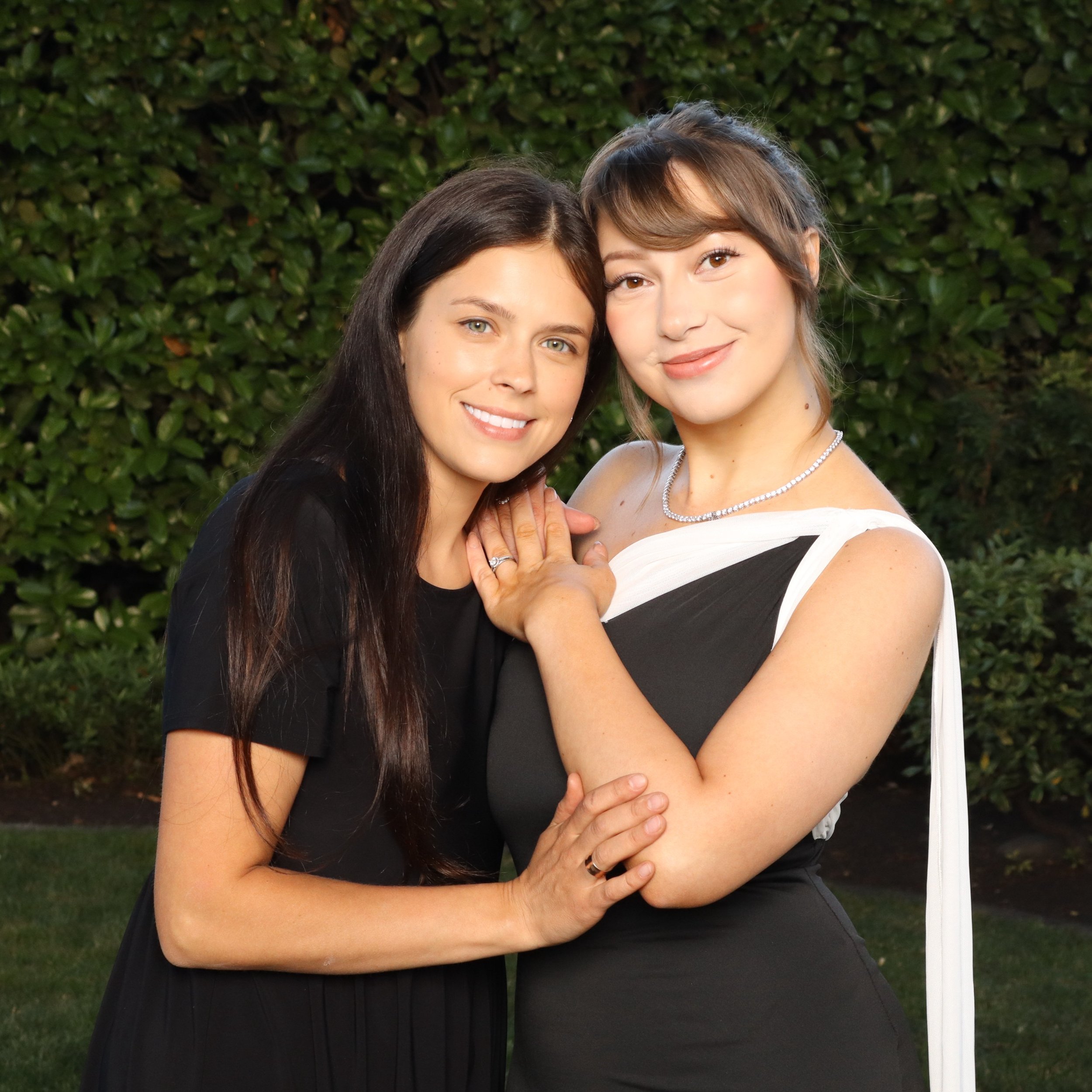 Two women standing close together, smiling, with greenery in the background.