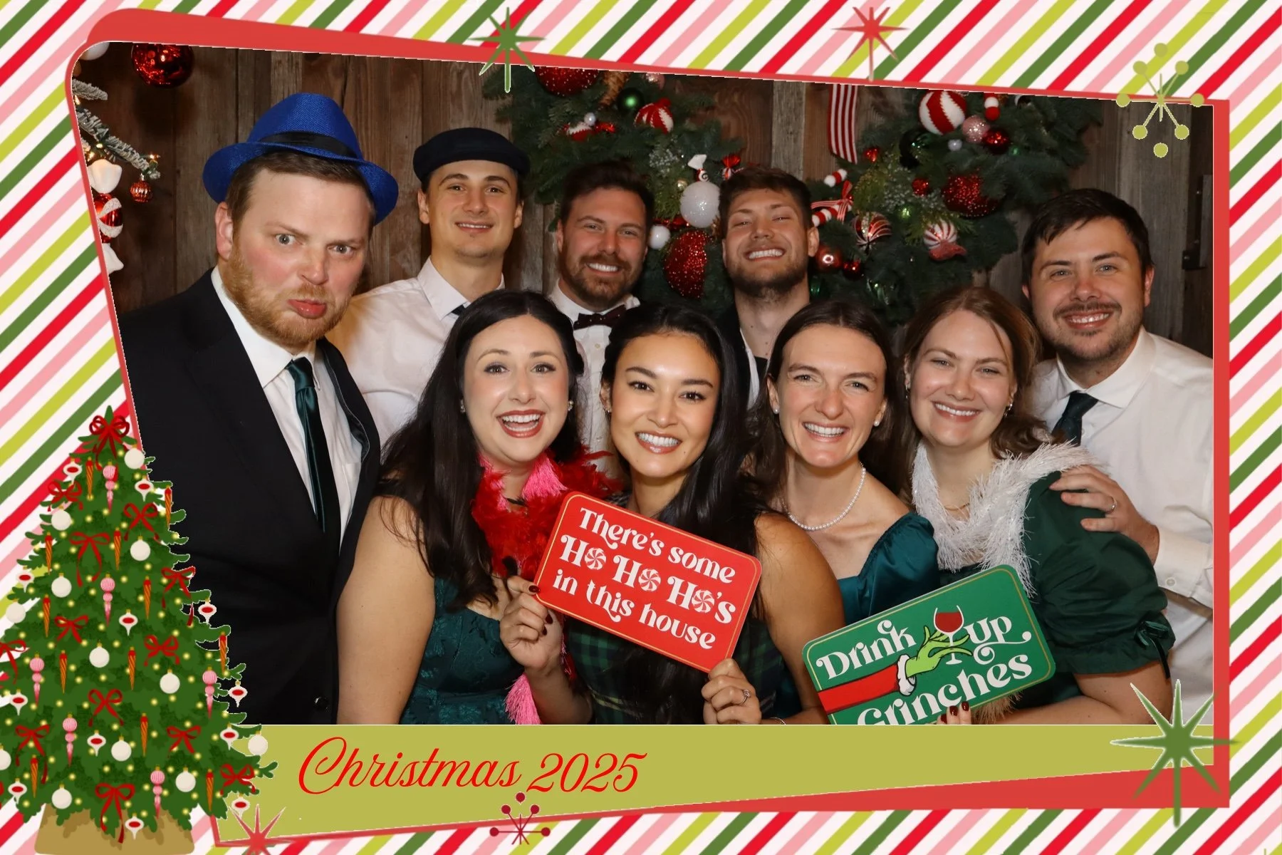 A group of people celebrating Christmas 2025, dressed festively, holding signs that say "There's some Ho Ho Ho in this house" and "Drink Up Ginchess", with Christmas trees and decorations in the background.