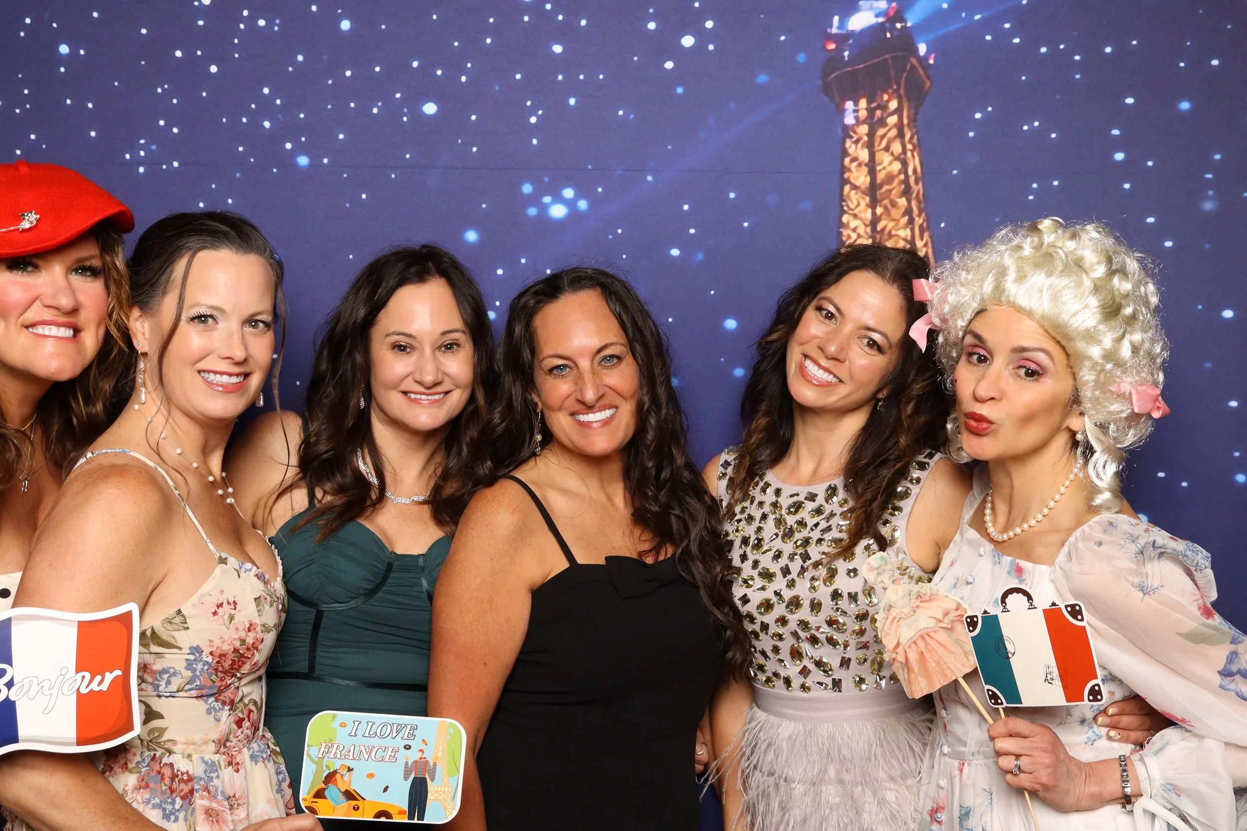 A group of six women smiling at a photo booth, with a backdrop of a lighthouse under a starry sky. Some women are holding signs with travel-themed messages, like 'Bonjour' and 'I Love France.' One woman is dressed in a white floral dress with a red hat, and another is wearing a curly blonde wig with pink ribbons.
