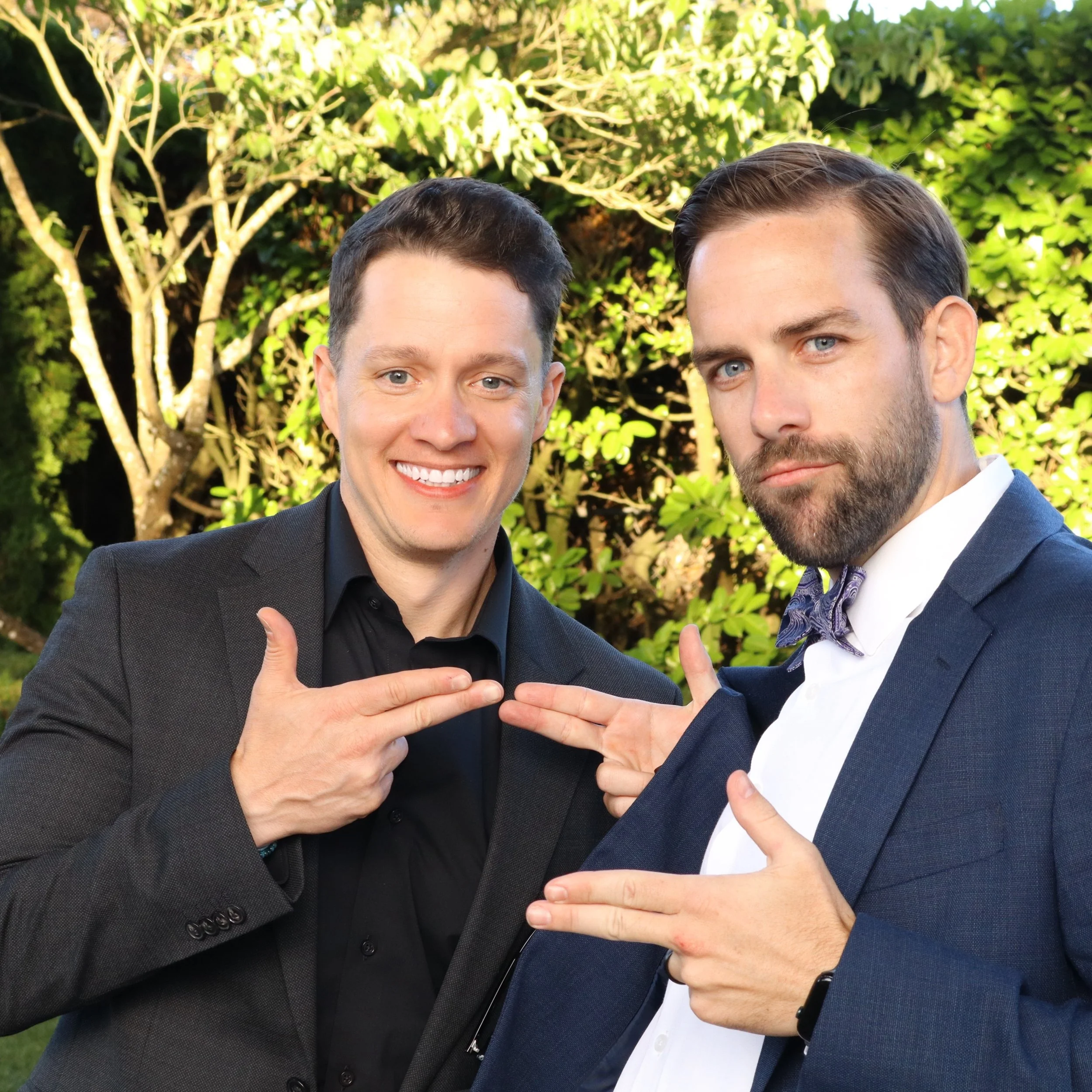 Two men in suits smiling and posing with their respective fists and palms facing each other in front of a green leafy background.