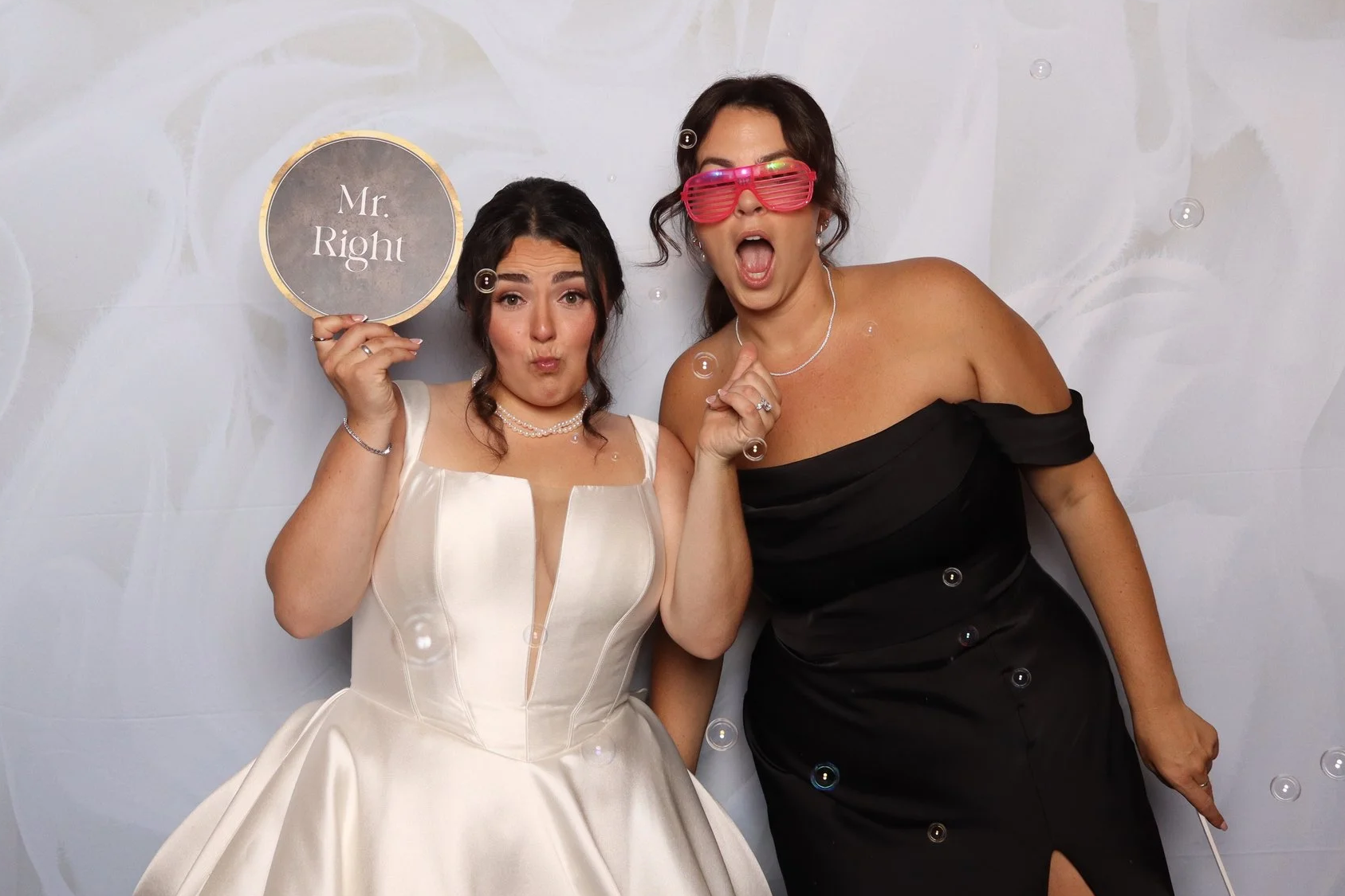 Two women dressed in formal attire, with one holding a round sign that says 'Mr. Right,' standing against a white backdrop with bubbles.