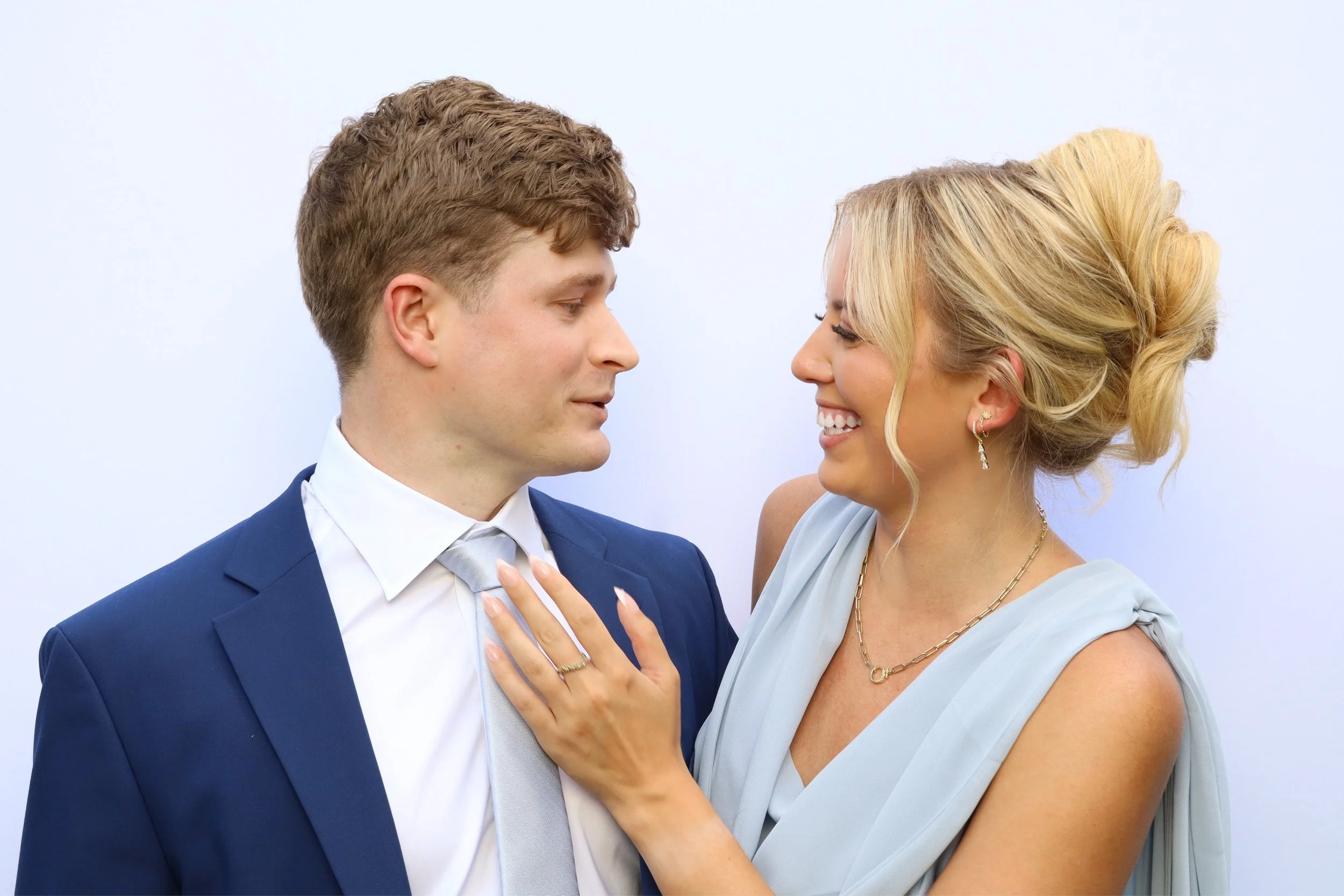 A man and woman dressed in formal attire facing each other, smiling, with the woman touching the man's chest.