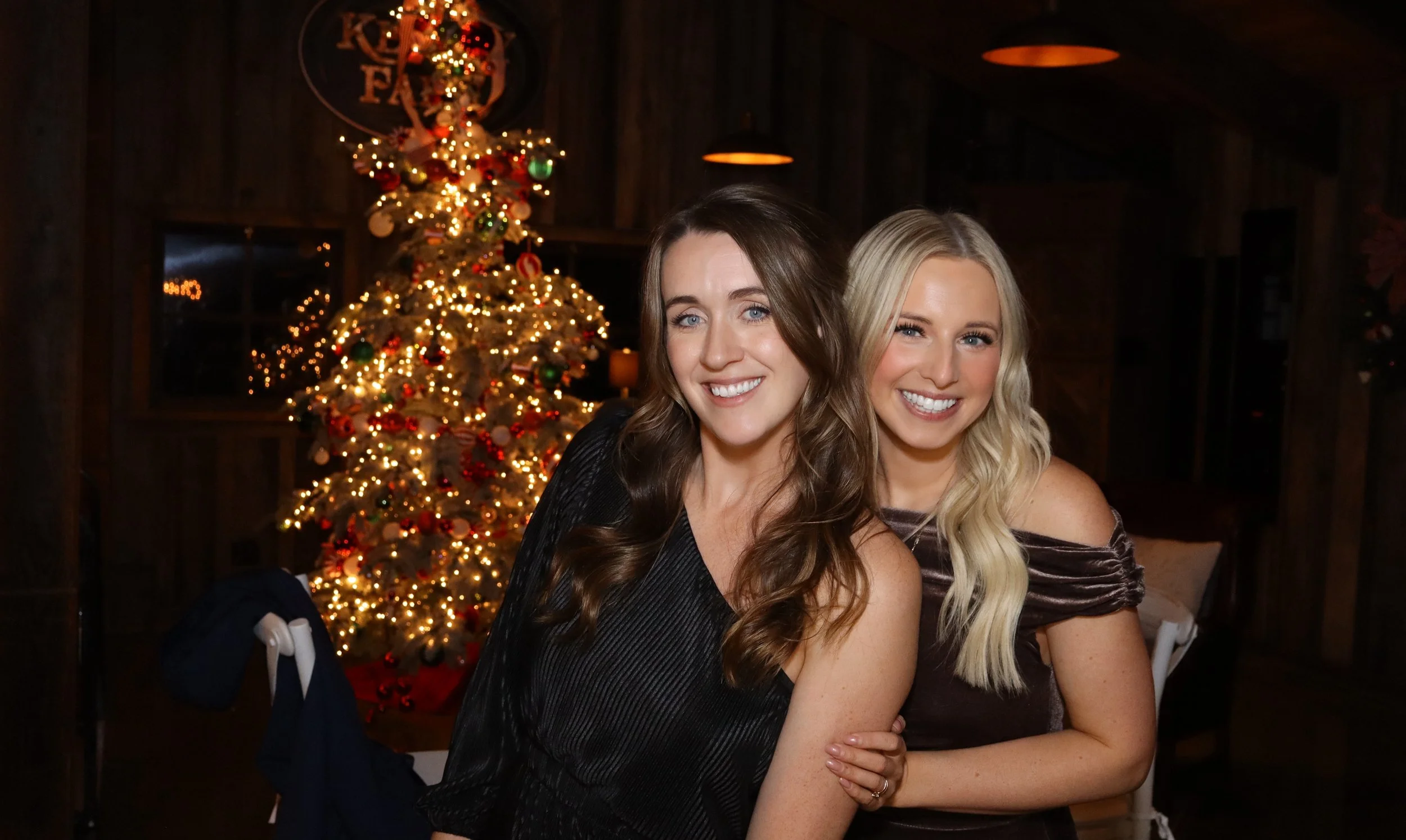 Two women smiling at a Christmas gathering, with a decorated Christmas tree with lights and ornaments in the background.