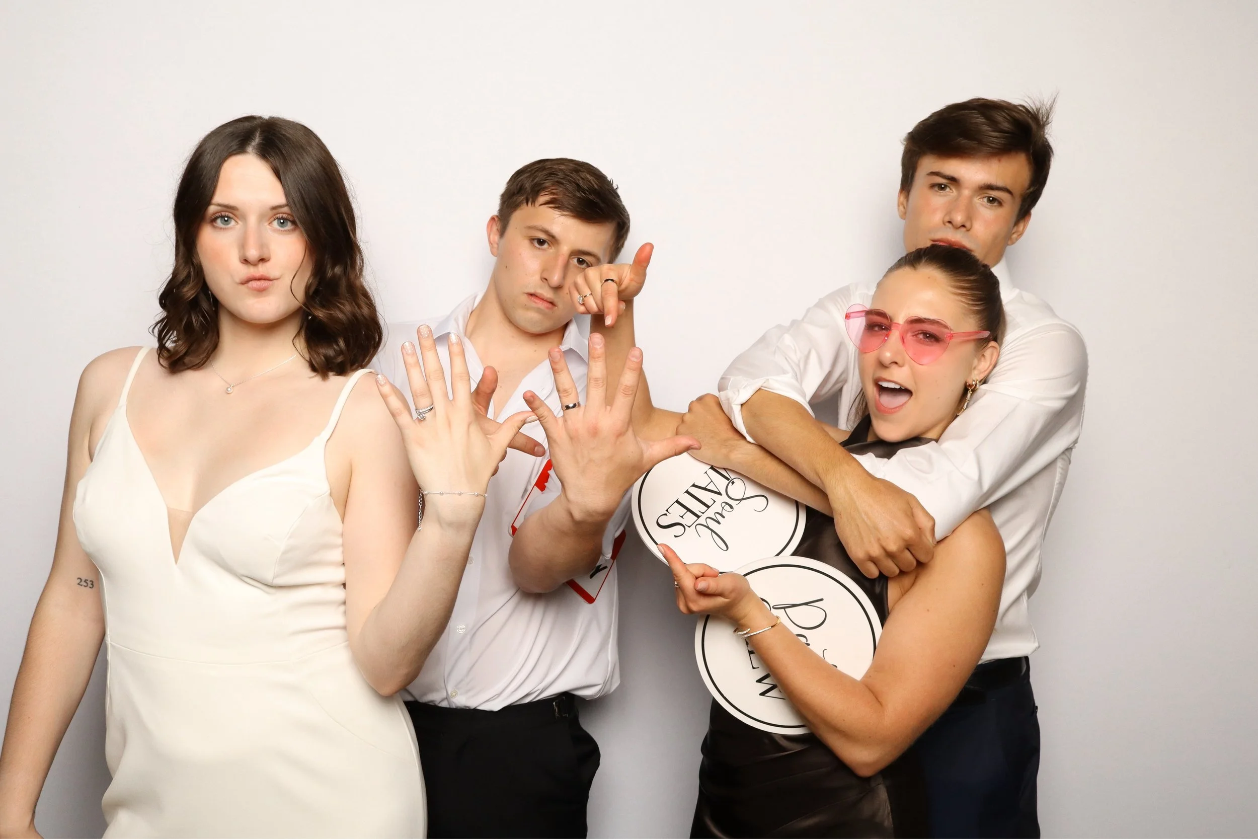Group of four young adults posing together against a plain white wall, holding round signs with text and making playful gestures.