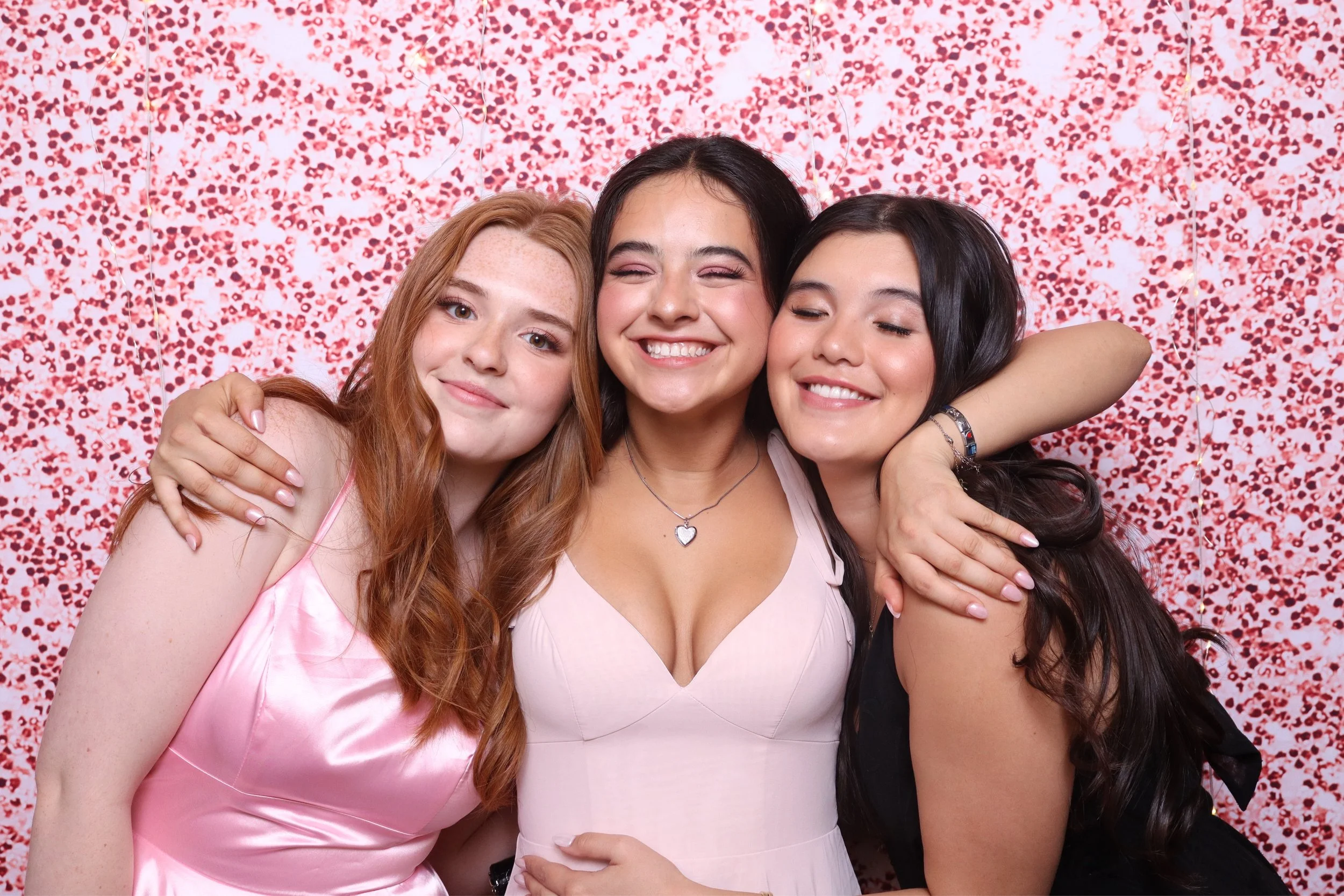 Three women hugging and smiling in front of a red and pink floral backdrop.