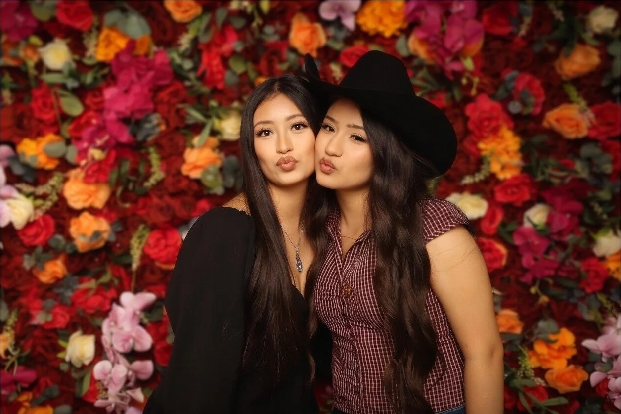 Two young women with long dark hair posing in front of a colorful floral backdrop, making kissy faces at the camera. One wears a black dress and a pendant necklace, the other wears a checkered top and a large black hat.