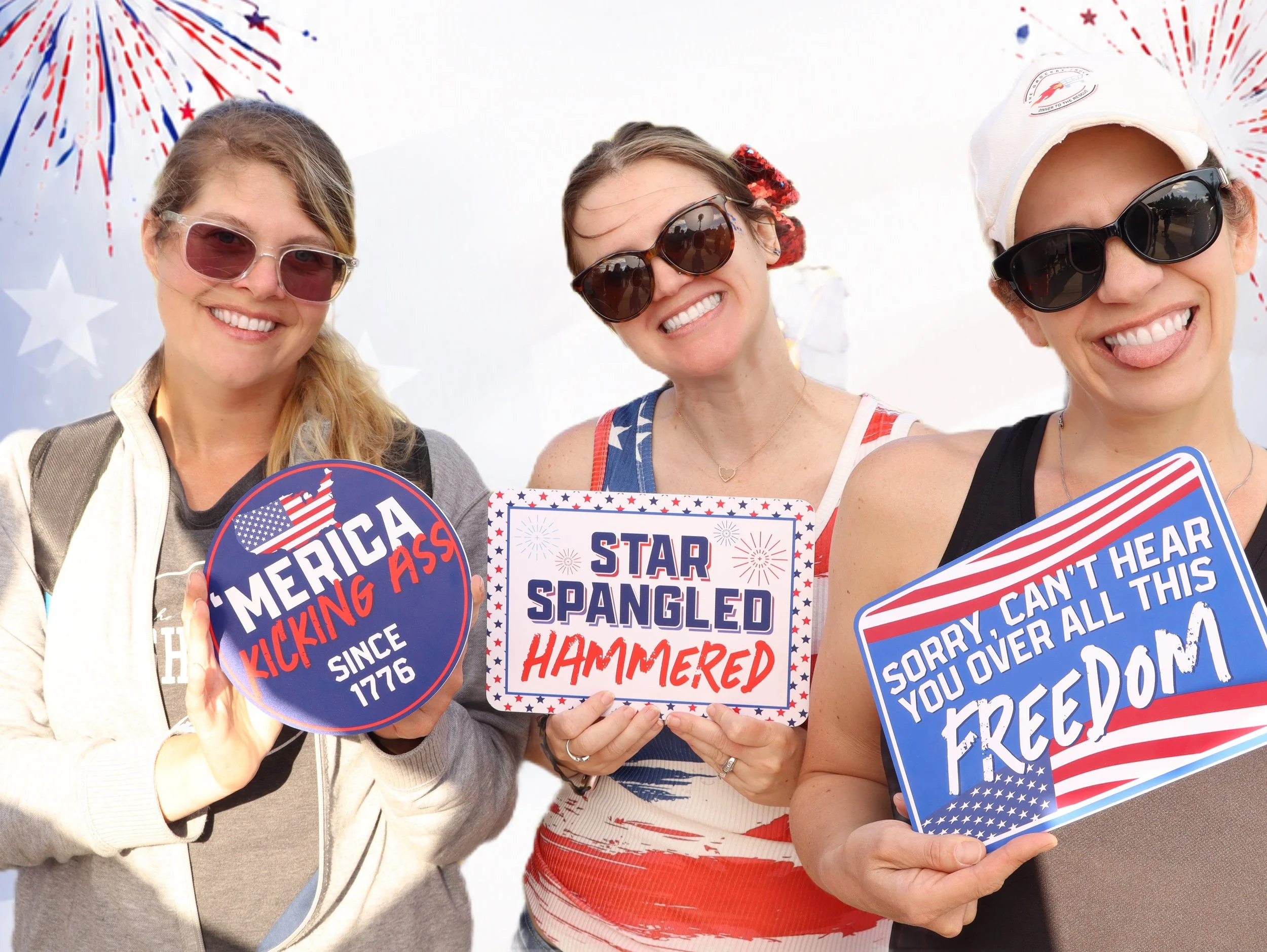 Three women happy and smiling, wearing sunglasses and holding patriotic signs with American themes, celebrating Independence Day with fireworks in the background.