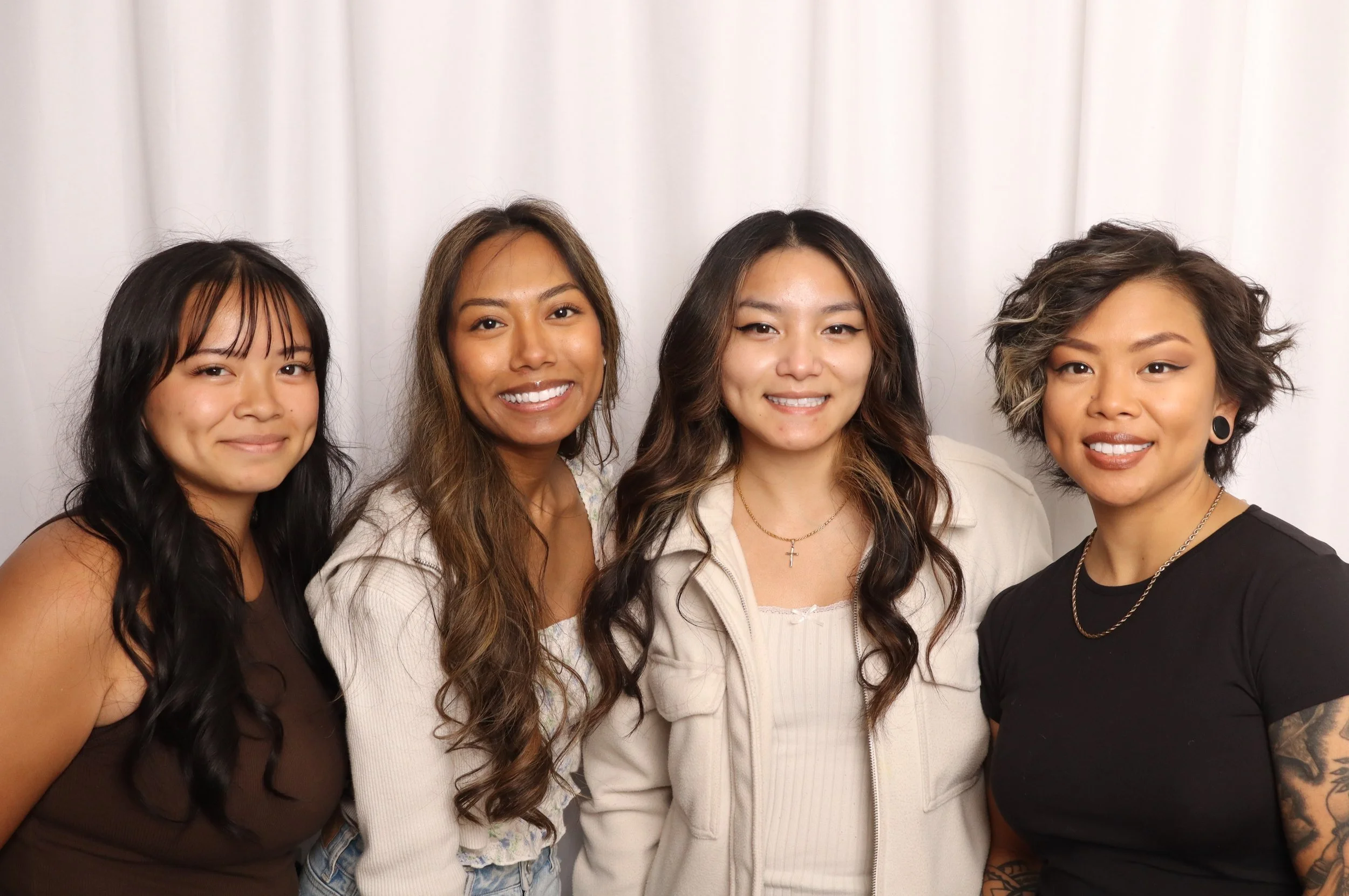 Group of four women smiling in front of a white curtain, with varied hairstyles and skin tones.