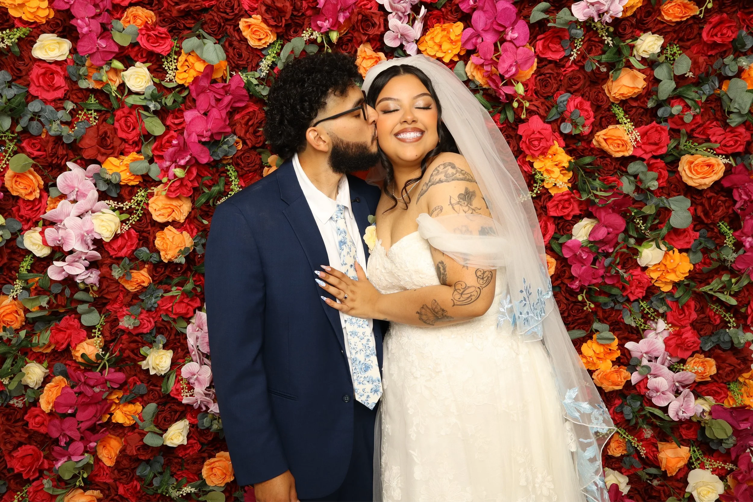A couple dressed in wedding attire, with the groom kissing the bride on her cheek, standing in front of a vibrant floral backdrop. The bride is smiling with her eyes closed, showing tattoos on her shoulder and arm. The groom is wearing glasses and a navy blue suit with a light-colored patterned tie. The background is filled with colorful red, pink, orange, yellow, and purple flowers and green leaves.