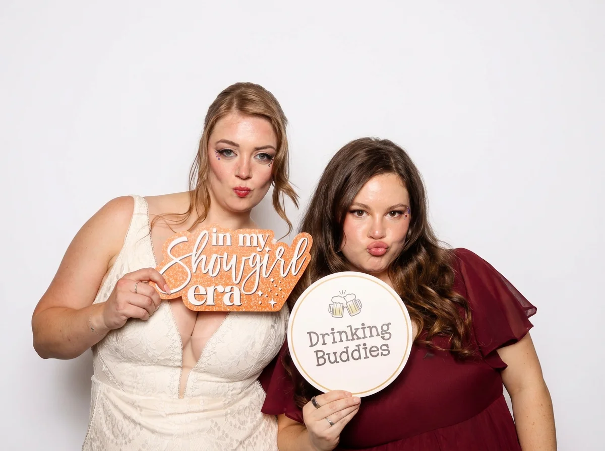 Two women posing together, one holding a sign that says 'in my showgirl era' and the other holding a sign that says 'Drinking Buddies'.