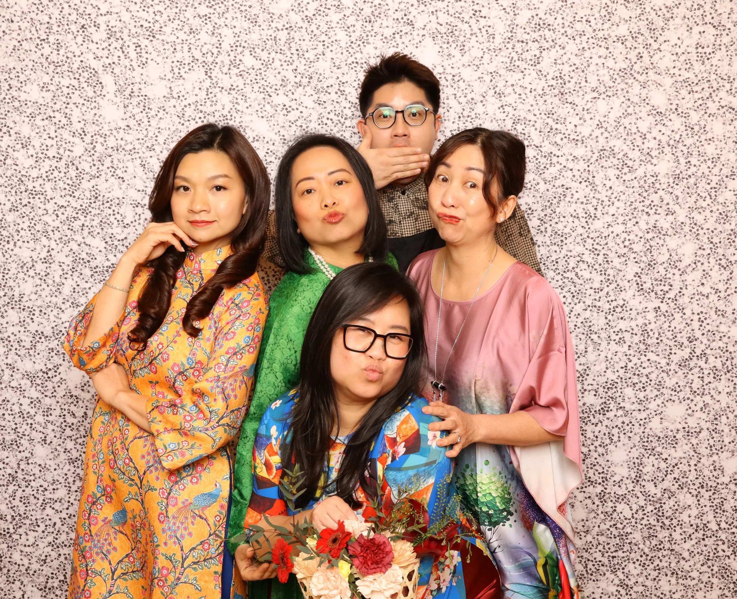 Group of five diverse women and one man posing for a photo against a speckled pink and gray background. The women are wearing colorful, floral and traditional clothing, some with floral jewelry. The man, positioned in the back, is wearing glasses and has his hand over his mouth in surprise. The woman in the foreground is holding a bouquet of red and cream flowers. Everyone appears to be enjoying themselves.