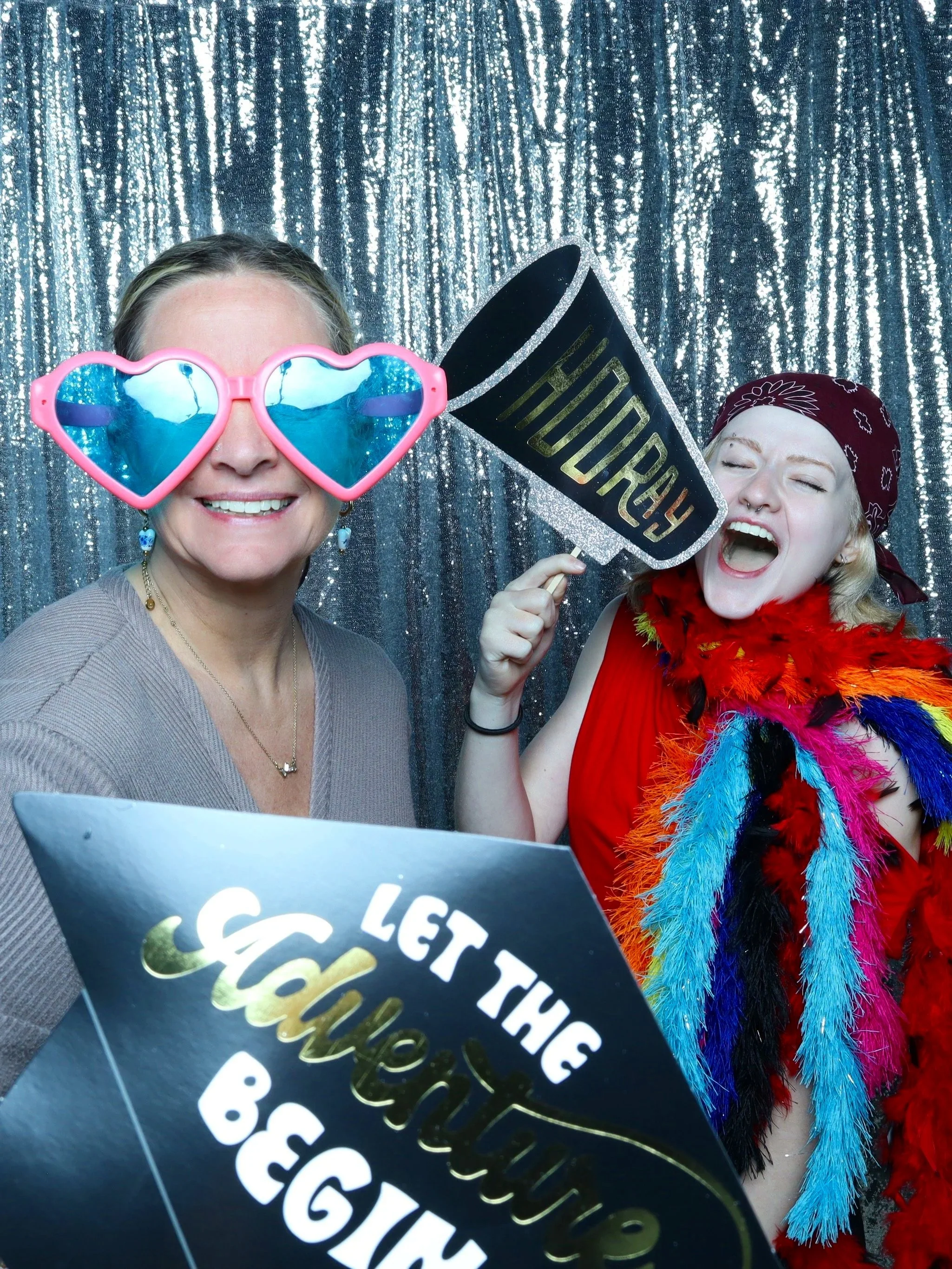 Two women are in front of a sparkly silver curtain. The woman on the left wears heart-shaped sunglasses with blue lenses and has earrings shaped like the state of Texas. The woman on the right is smiling widely, has a nose ring, and is wearing a bandana with a floral pattern, a red top, and a colorful feathered boa. She holds a black sign with the word 'HURRY' in gold lettering. They appear to be at a photo booth or celebration.