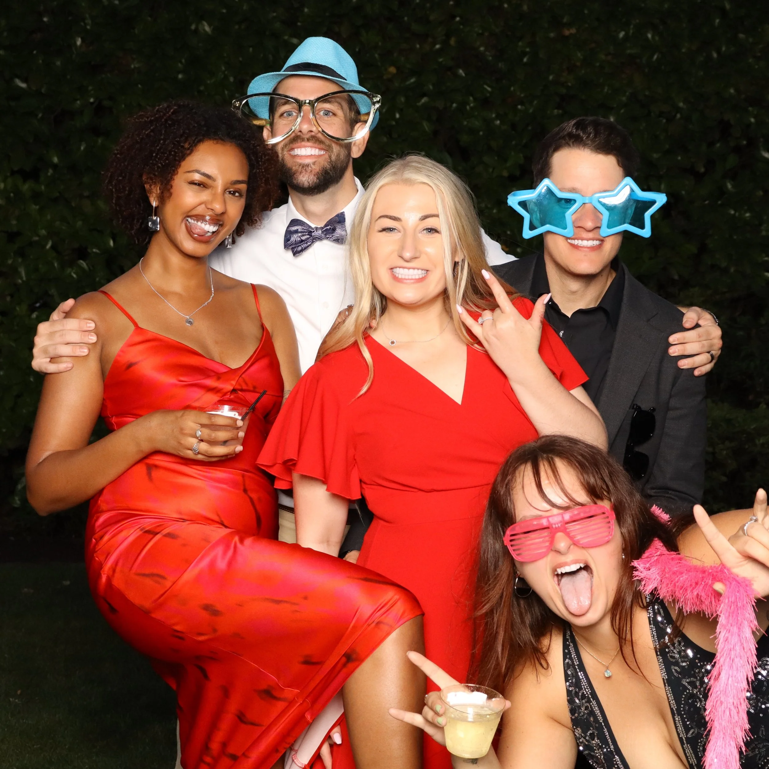 Group of four people at a party outdoors, smiling and posing for the camera. Two women and two men wearing festive accessories, including star-shaped sunglasses, a blue hat with large glasses, and holding drinks. A woman in a red dress is making a pe