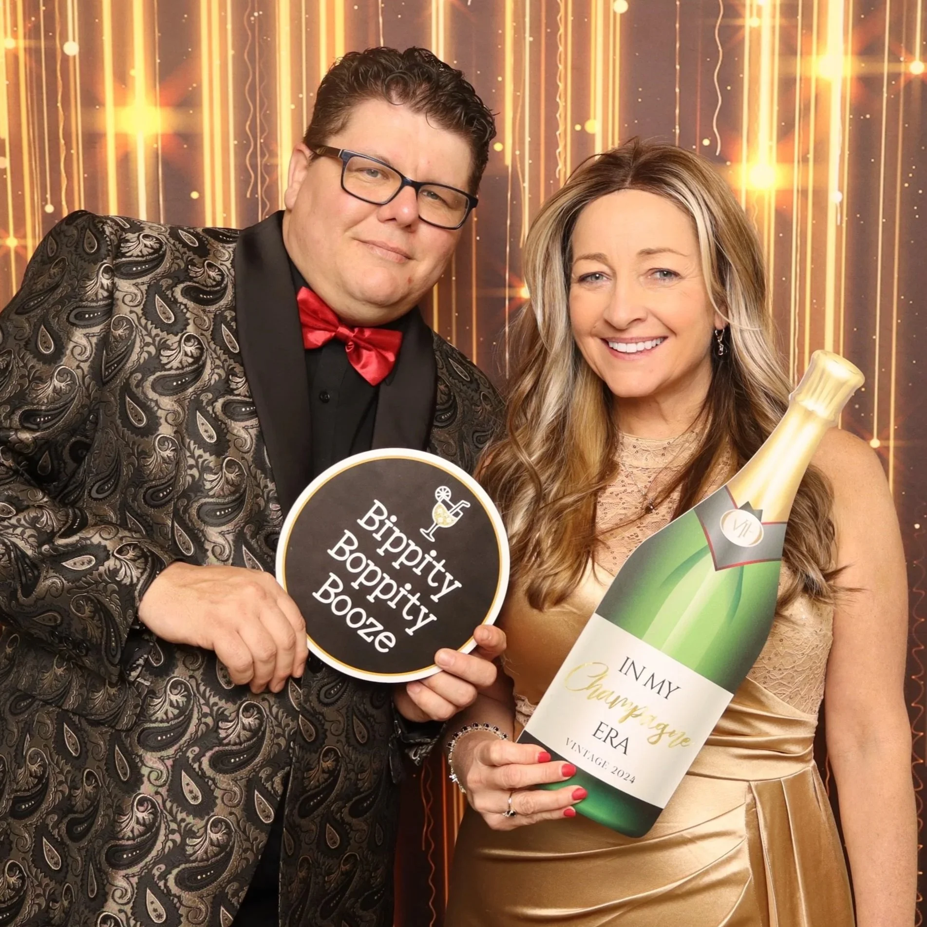 A man and woman dressed in formal attire smiling at a celebration event. The man is wearing a paisley patterned jacket, black shirt, and red bow tie, and is holding a sign that says 'Hippity Boppity Booze.' The woman has long blonde hair, is wearing a gold dress, and is holding a windowed bottle and a card with a thank you message. The background has vertical golden streamers with lights.