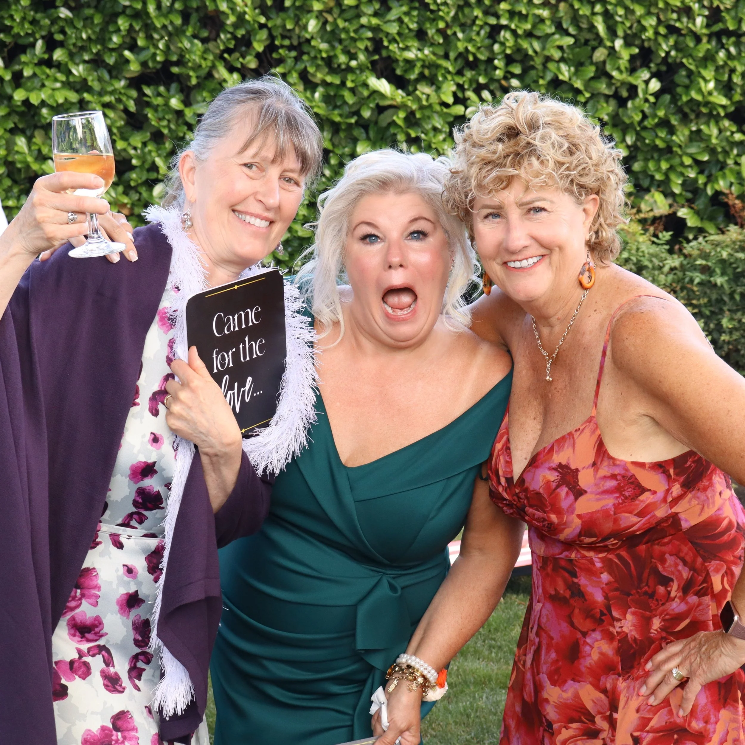 A group of four women smiling and celebrating outdoors, with some holding a glass of rosé wine and a sign that says 'Came for the'. They are dressed in colorful clothing, and there is a green hedge in the background.