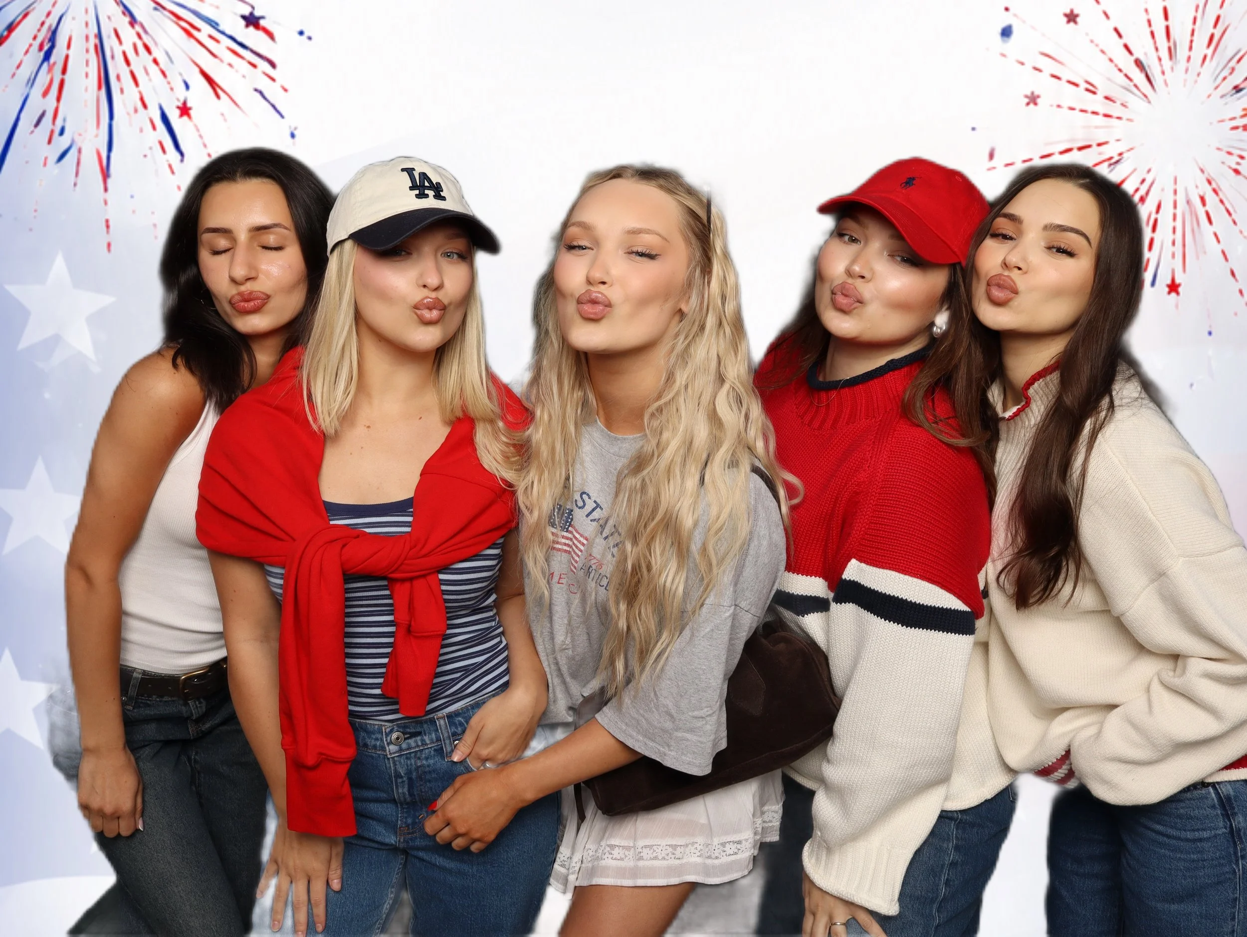 A group of five young women posing together with a patriotic theme, with fireworks and stars in the background. They are wearing casual clothes and hats, with some making kissing faces.