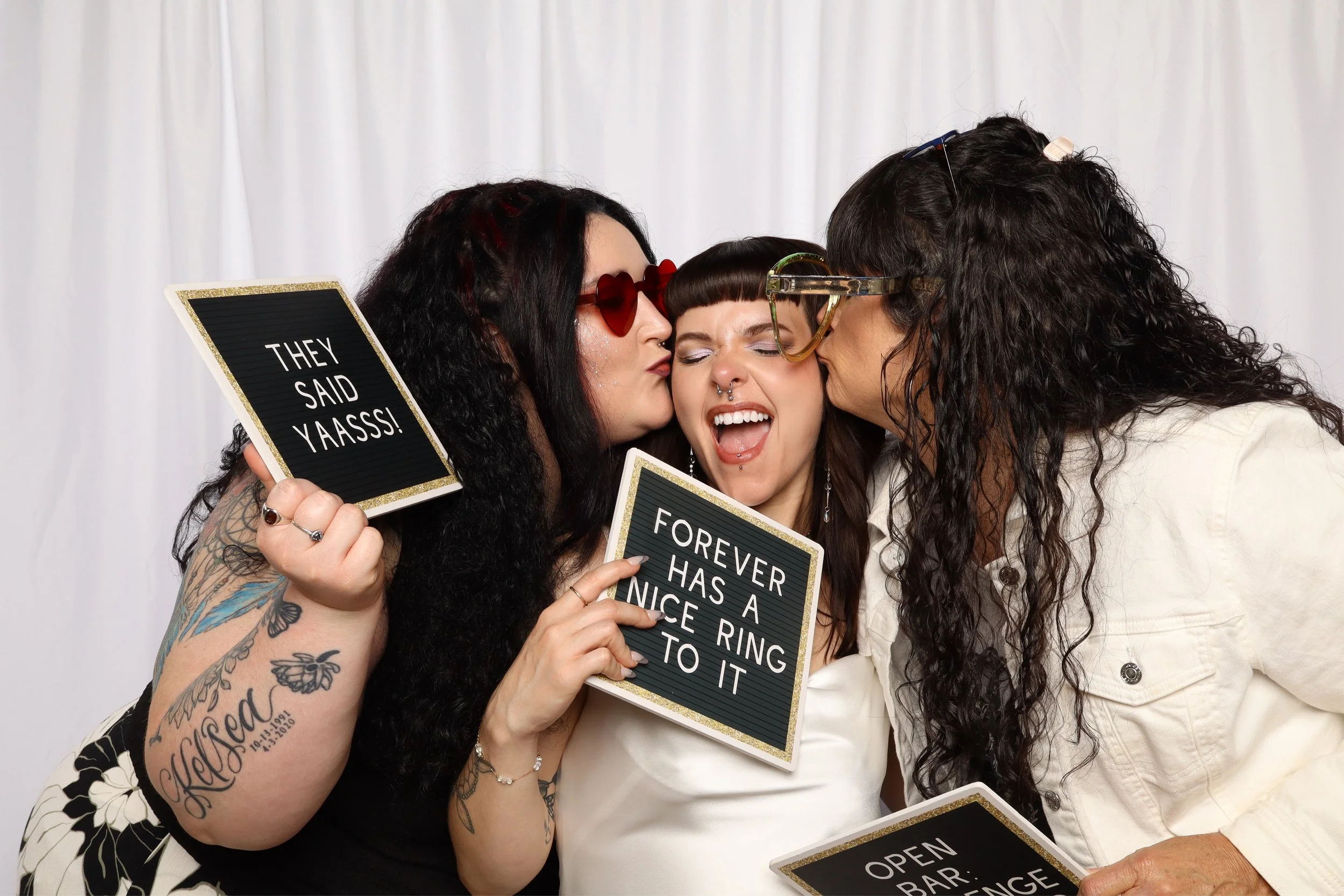 Three women are kissing and hugging each other, holding signs with messages like "They said YAASS!" and "Forever has a nice ring to it." They are smiling and seem happy, with a white backdrop behind them.