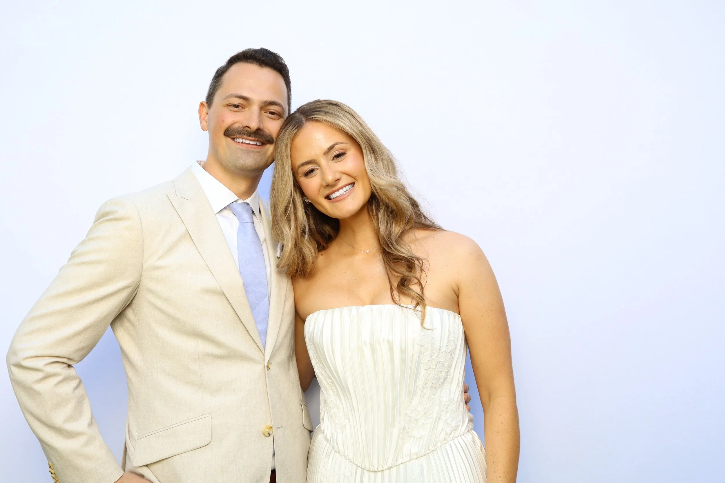 A smiling man with dark hair and a mustache wearing a light-colored suit and a woman with long blonde hair wearing a strapless light-colored dress pose together against a plain white background.
