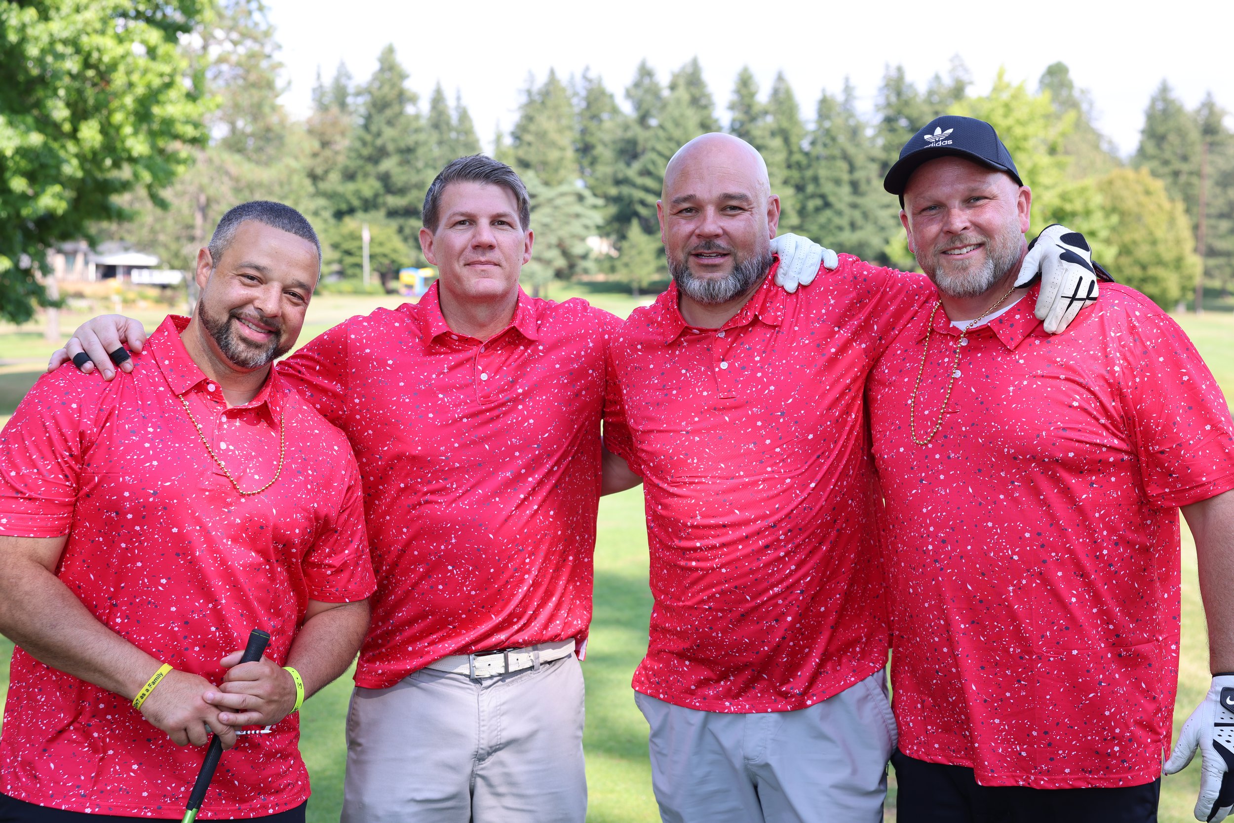 Four men standing together on a golf course, wearing matching red shirts with white and blue splatter patterns, smiling. One is holding a golf club, and another has golf gloves, with a background of trees and a sunny sky.