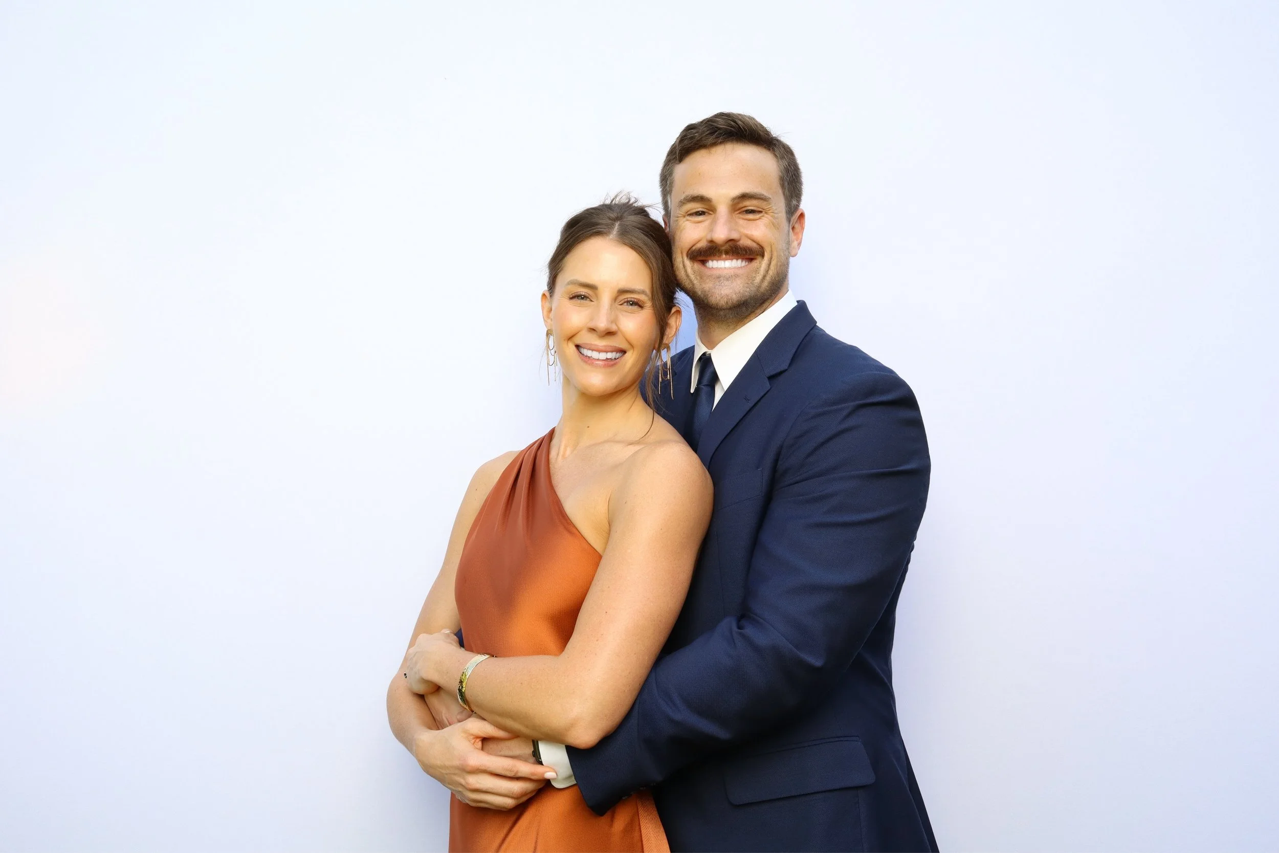 A smiling man and woman stand closely together against a plain white background, with the man wearing a dark blue suit and the woman in a rust-colored dress.