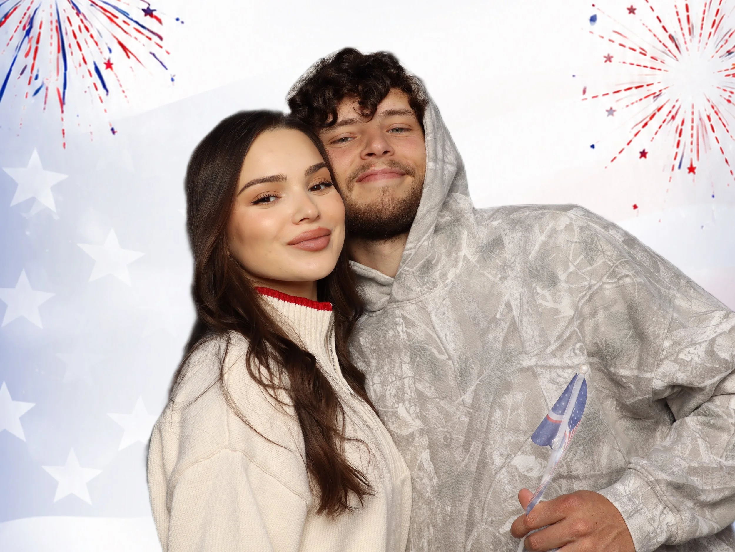 A young woman with long brown hair and a young man with curly hair and a beard taking a selfie together at a celebration. The background features a white backdrop with blue and red fireworks and star patterns, and the man is holding a small confetti 