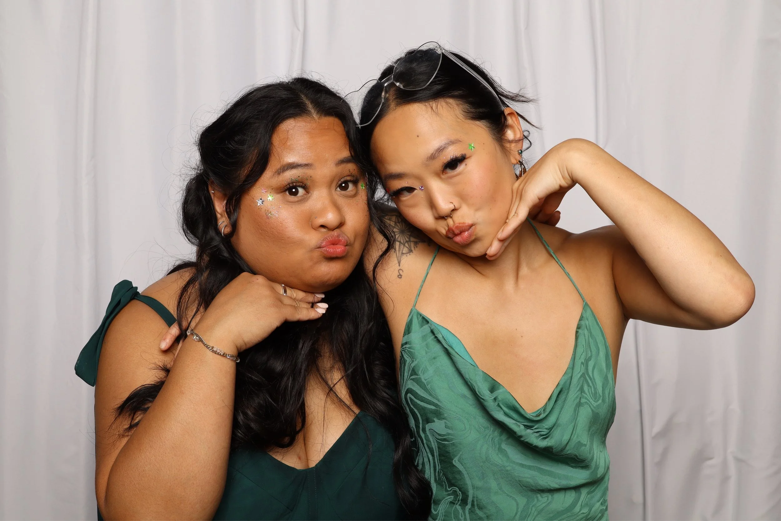 Two women posing close together, one with dark wavy hair and the other with short dark hair, both wearing green dresses, decorated with colorful star-shaped confetti and stickers, in front of a plain white backdrop.
