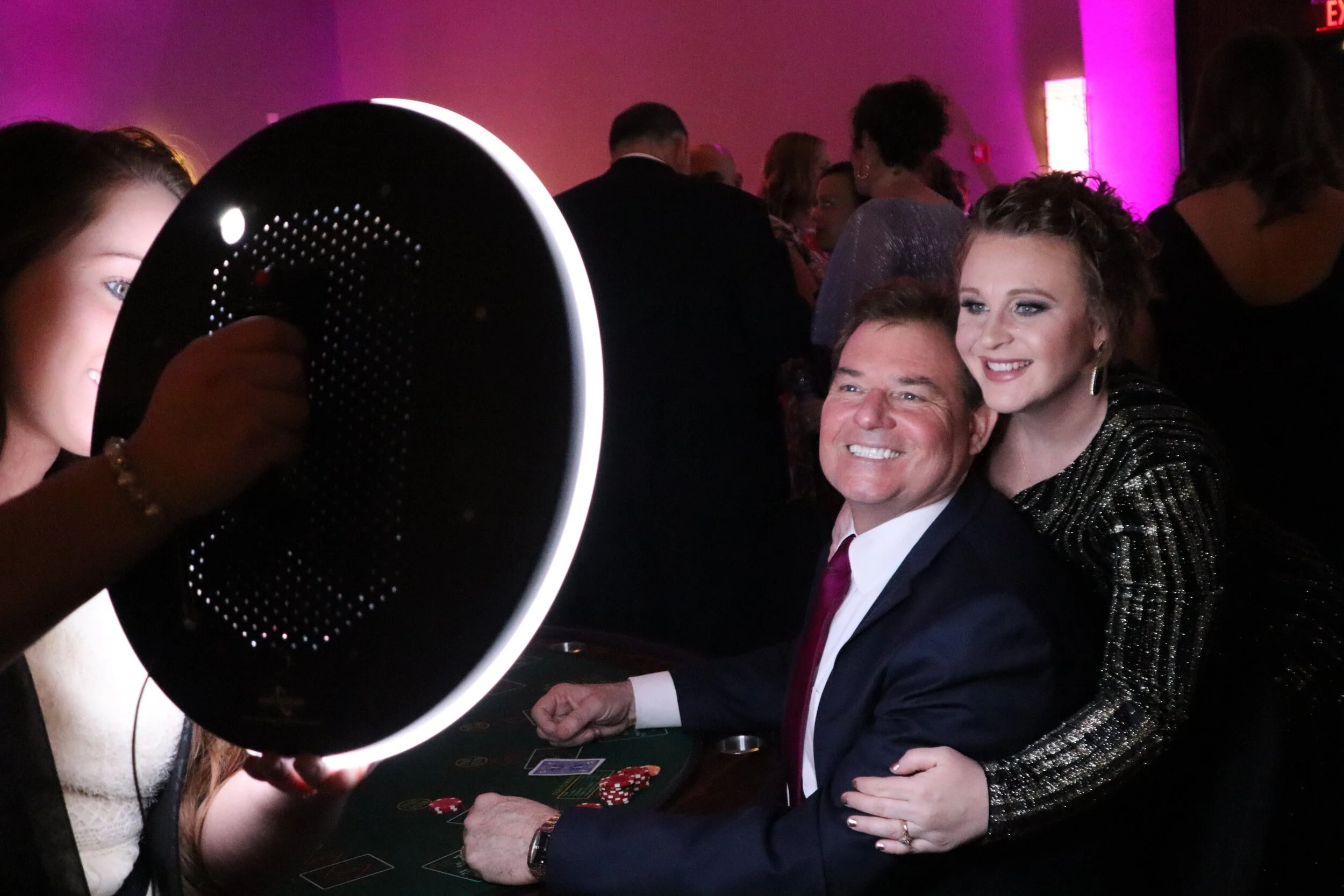 People playing poker at a casino table with a large circular lamp overhead, in a dimly lit environment with pink and purple lighting.