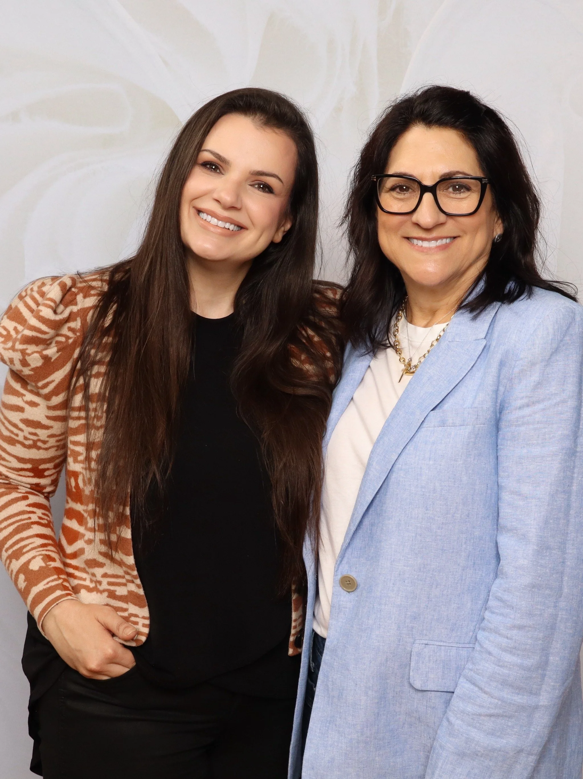 Two women smiling, one with long brown hair, wearing a black top and a patterned blazer, the other with shoulder-length dark brown hair, wearing glasses, a light blue blazer, and a white top.