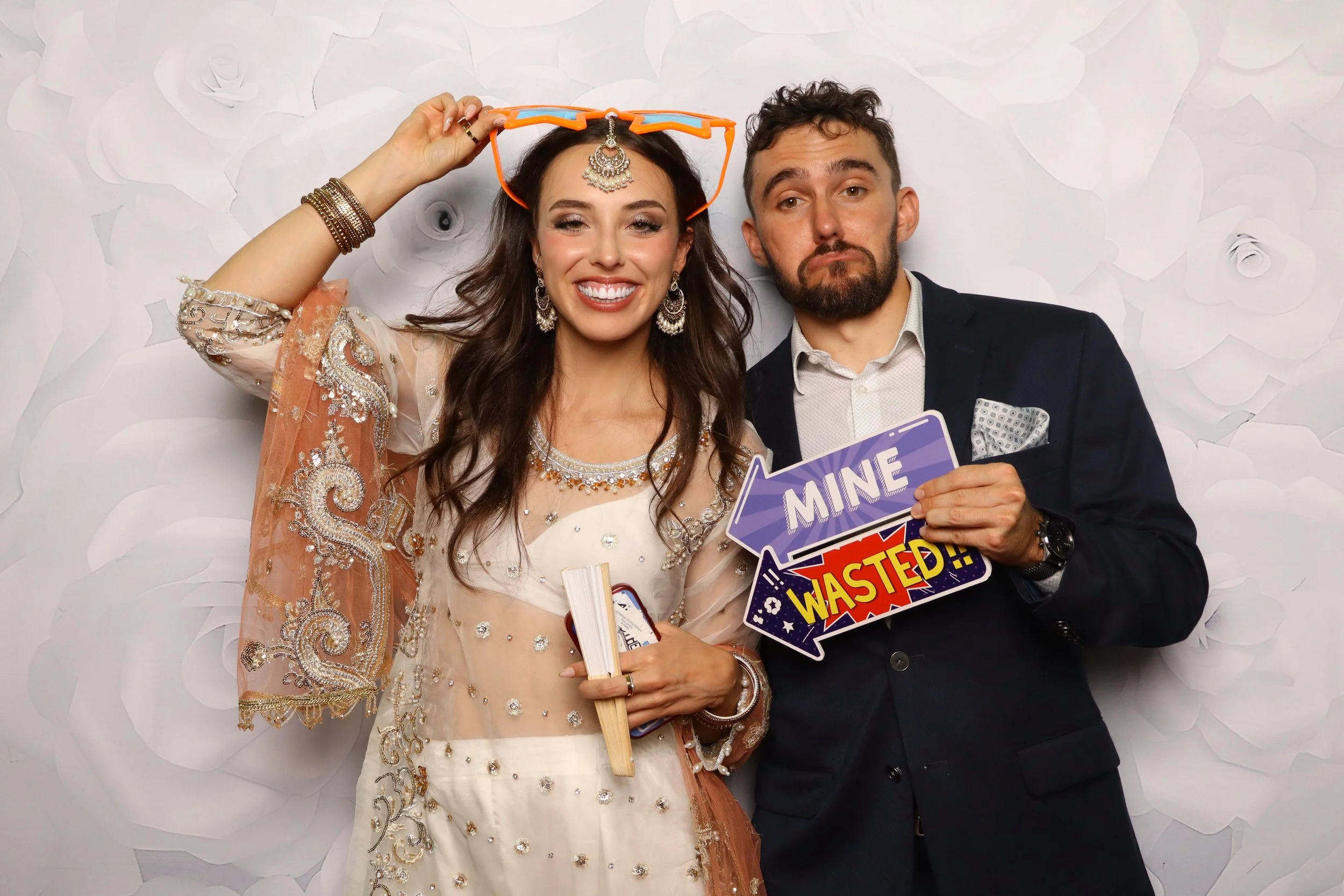 A smiling woman in a traditional outfit holding a sign that says 'WASTED,' with a man standing next to her holding a sign that says 'MINE.' They are in front of a white textured wall.
