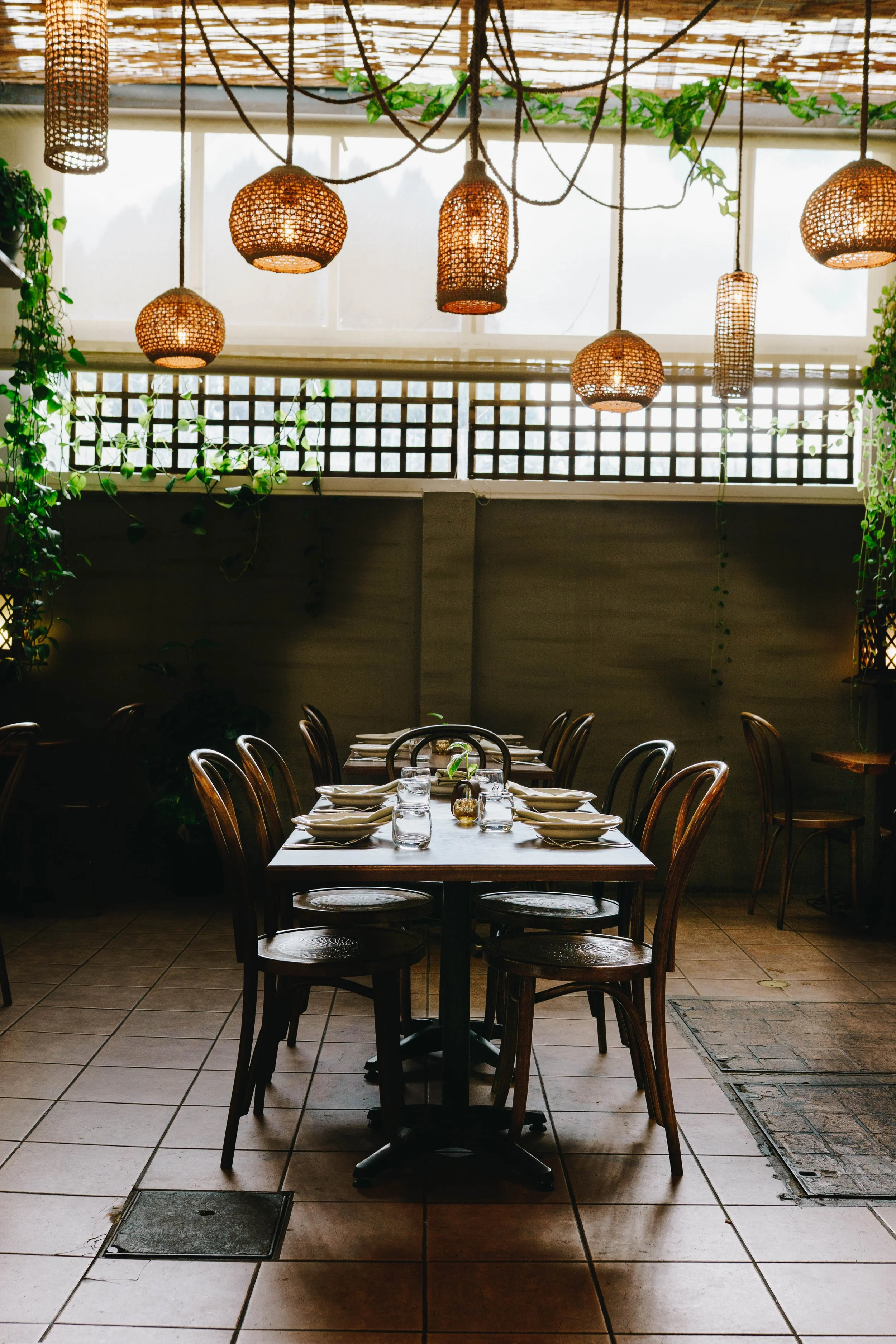A restaurant dining area with a long wooden table set with plates, glasses, and napkins. Surrounding chairs are wooden with curved backs. Light filters through woven pendant lamps hanging from the ceiling, with greenery and latticework visible in the background.