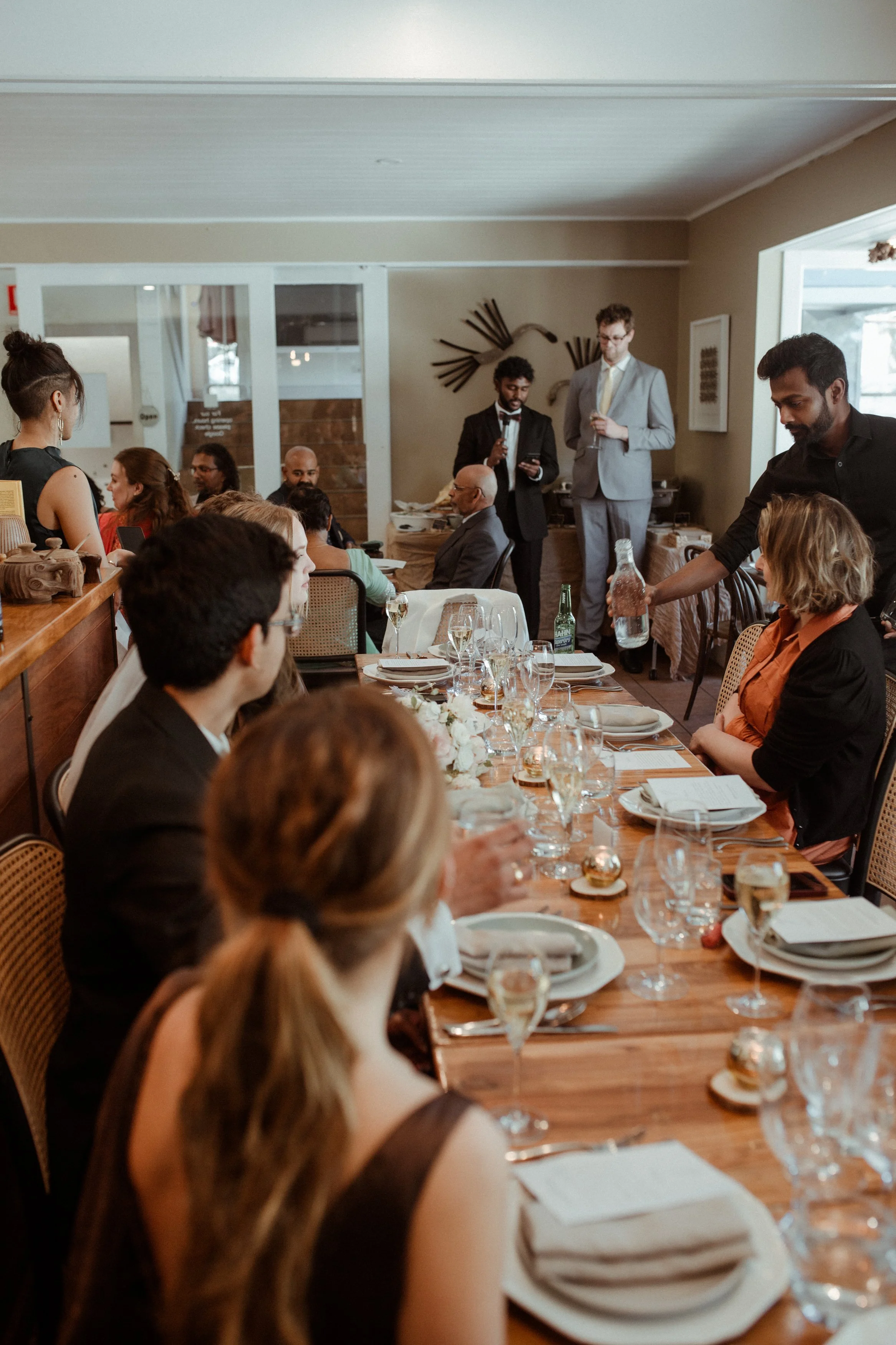 People gathered around a long dining table with wine glasses, plates, and a floral centerpiece, in a warmly decorated restaurant or event space, with some standing and some seated, engaging in conversation.