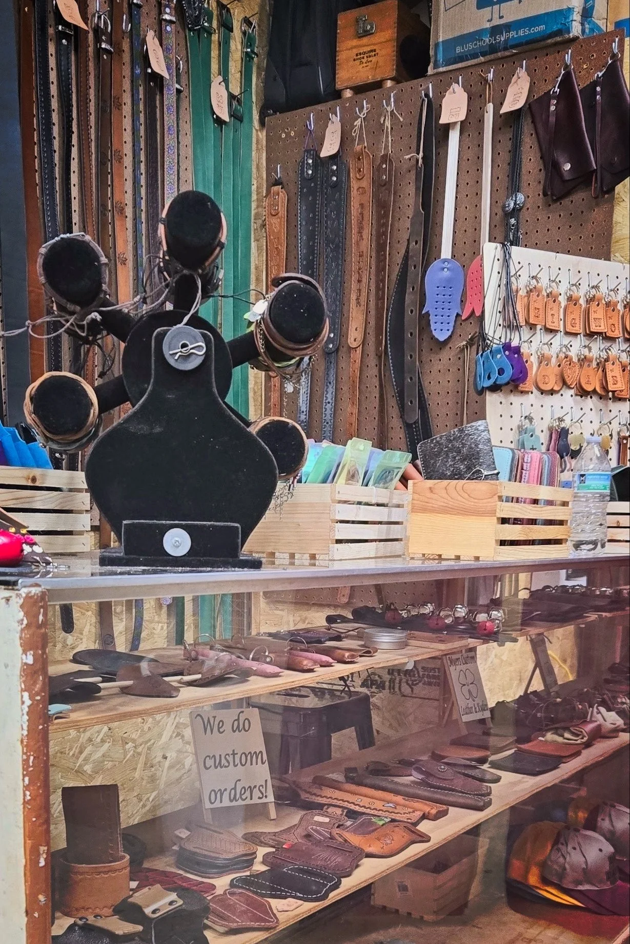 A display of leather belts, wallets, and keychains in a store with a wooden pegboard background. There are various accessories and signs indicating customization options.