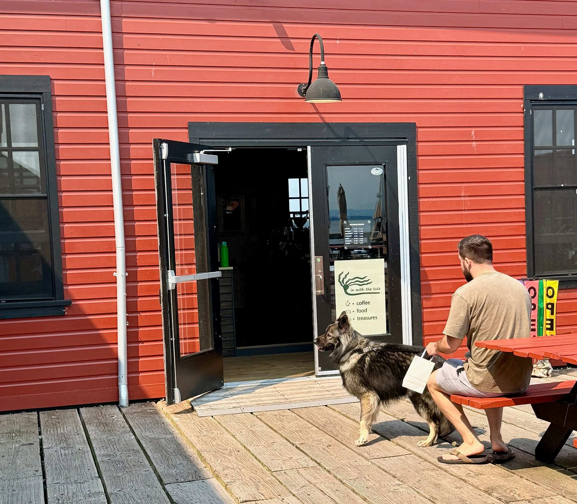 A man sitting on a red wooden bench outdoors, accompanied by a large, fluffy dog at his side. They're in front of a red building with black window frames and an open black door leading inside. The man is holding a shopping bag, and there's a sign nea