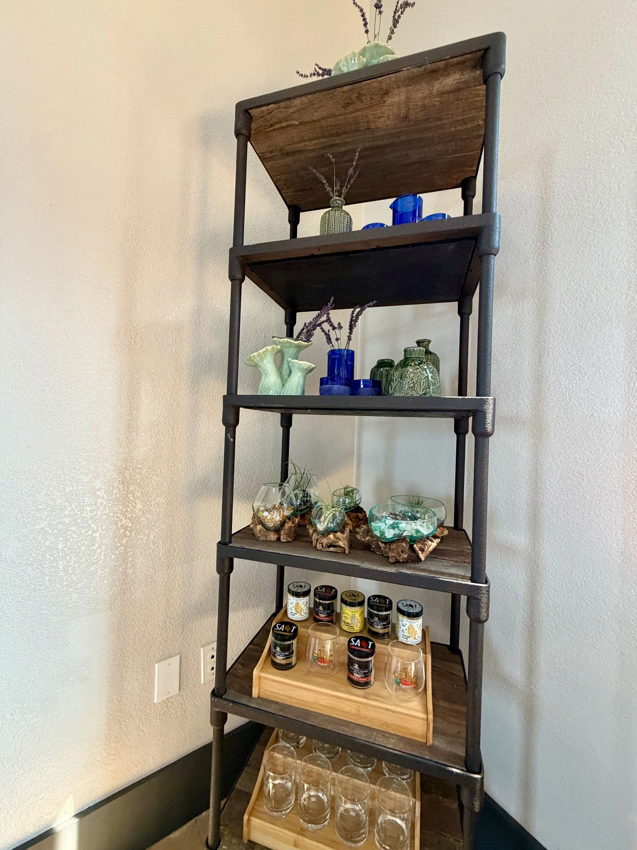 A five-tiered shelving unit with decorative items and containers, including vases, jars, and glasses, against a plain wall.
