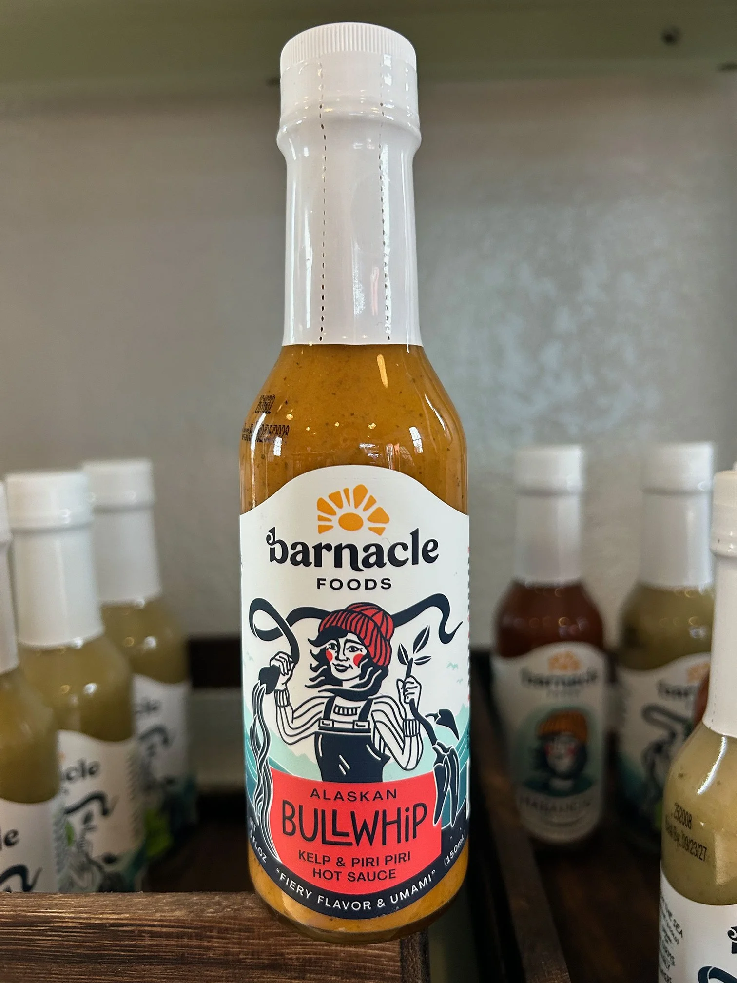 A bottle of Alaskan Bullwhip hot sauce with a colorful label featuring a cartoon woman with red hair, holding a chili pepper, and the text indicating it is flavored with jalapeño, Fresno, and chili. The background shows other bottles of hot sauce on 