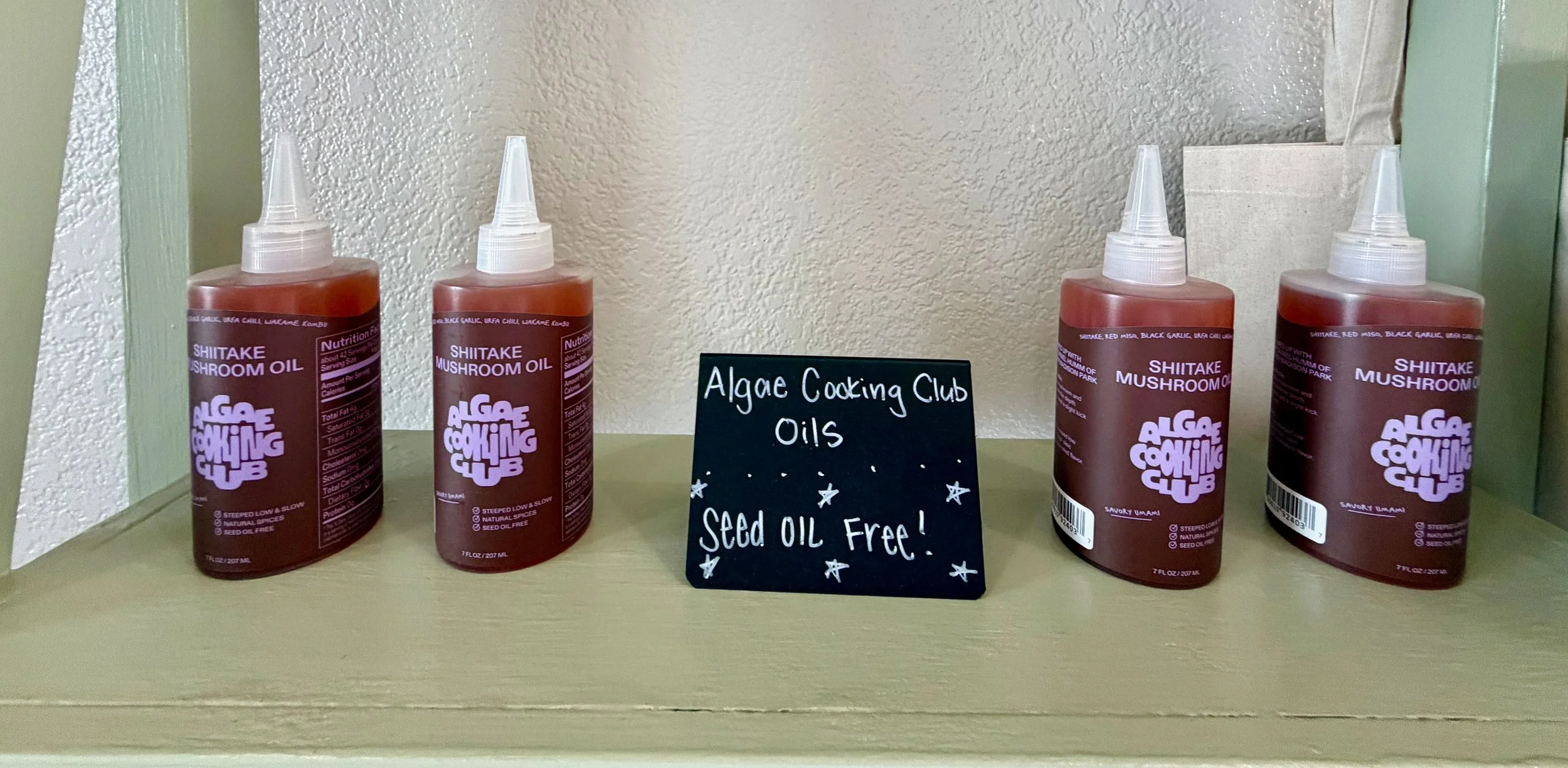 Four bottles of Shiitake mushroom oil from Algor Cooking Club displayed on a shelf with a sign that reads 'Algae Cooking Club Oils. Seed oil free!'